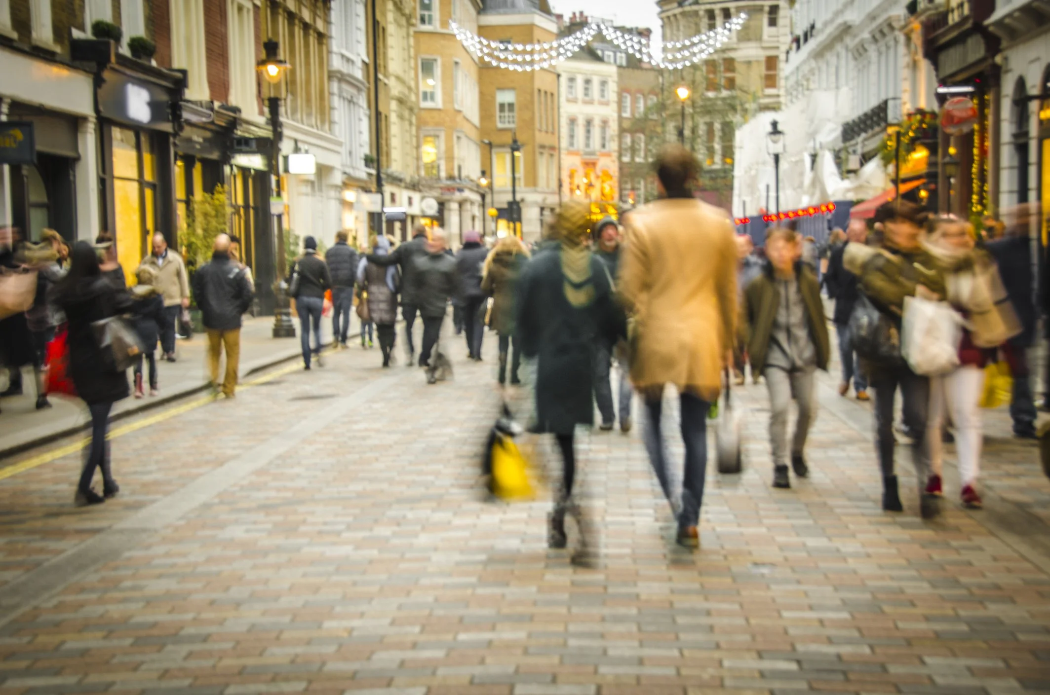 A busy pedestrian street with people walking and shopping, decorated with string lights overhead, lined with shops and cafes, with historic buildings in the background.