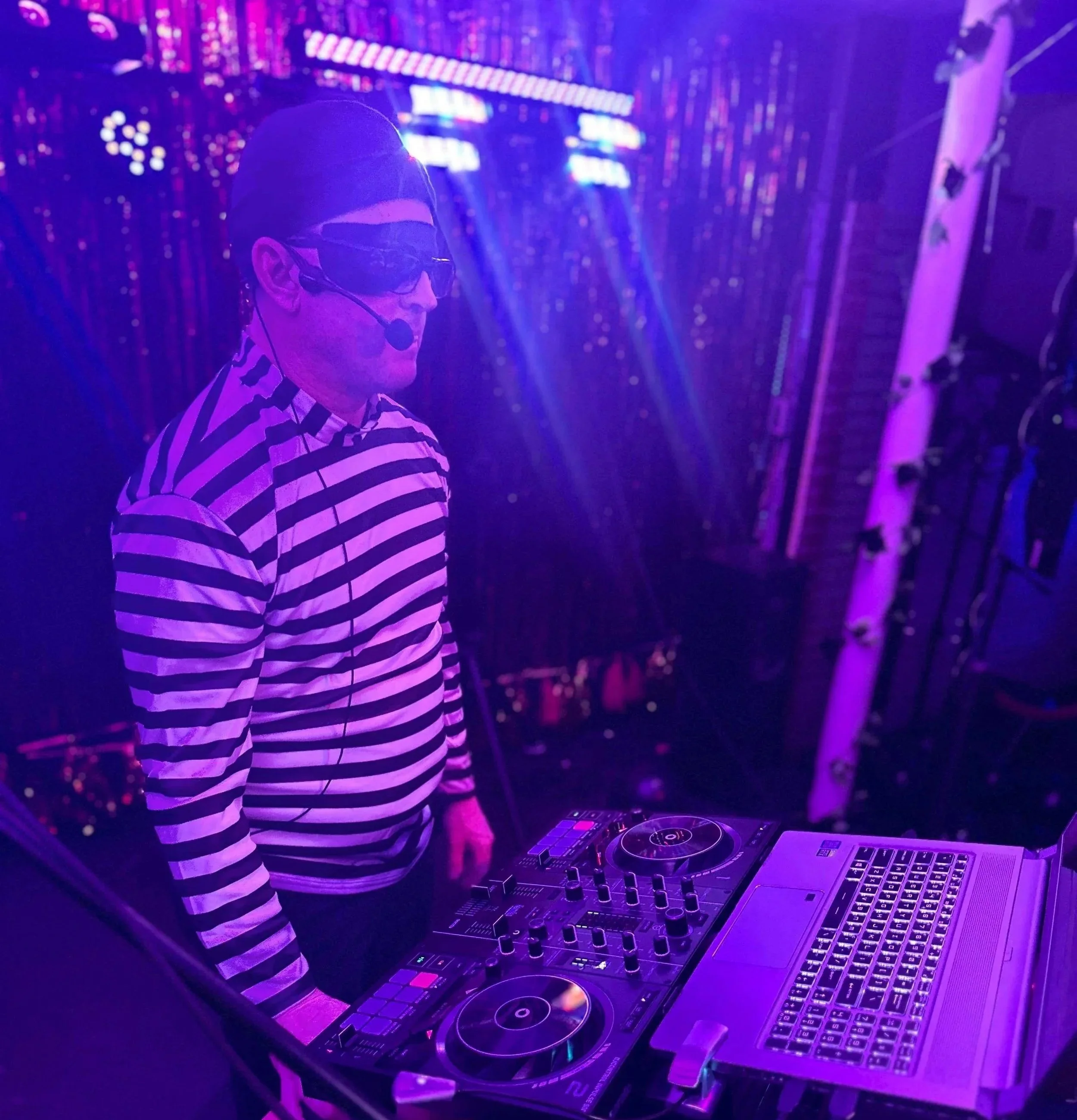 DJ performing at a nightclub with purple lighting, wearing sunglasses and a striped shirt, behind DJ equipment including turntables and a laptop.
