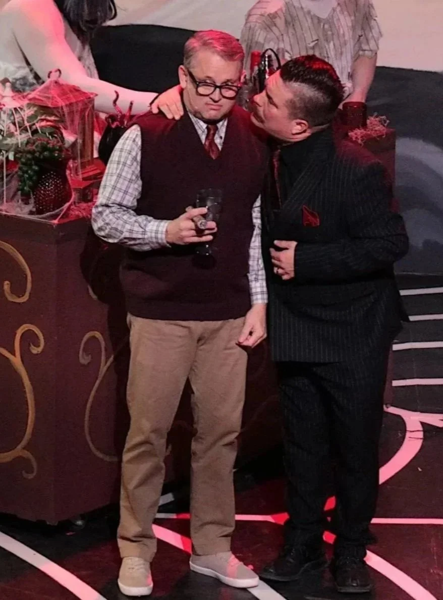 Two men in formal attire are standing close together on stage. One man is holding a glass of drink and has an amused, skeptical expression, while the other man is leaning in and speaking to him. There are people in the background.
