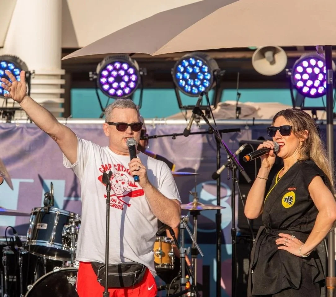 Two people speaking or singing into microphones on a stage with colorful lights and a drum set in the background. Both are wearing sunglasses.