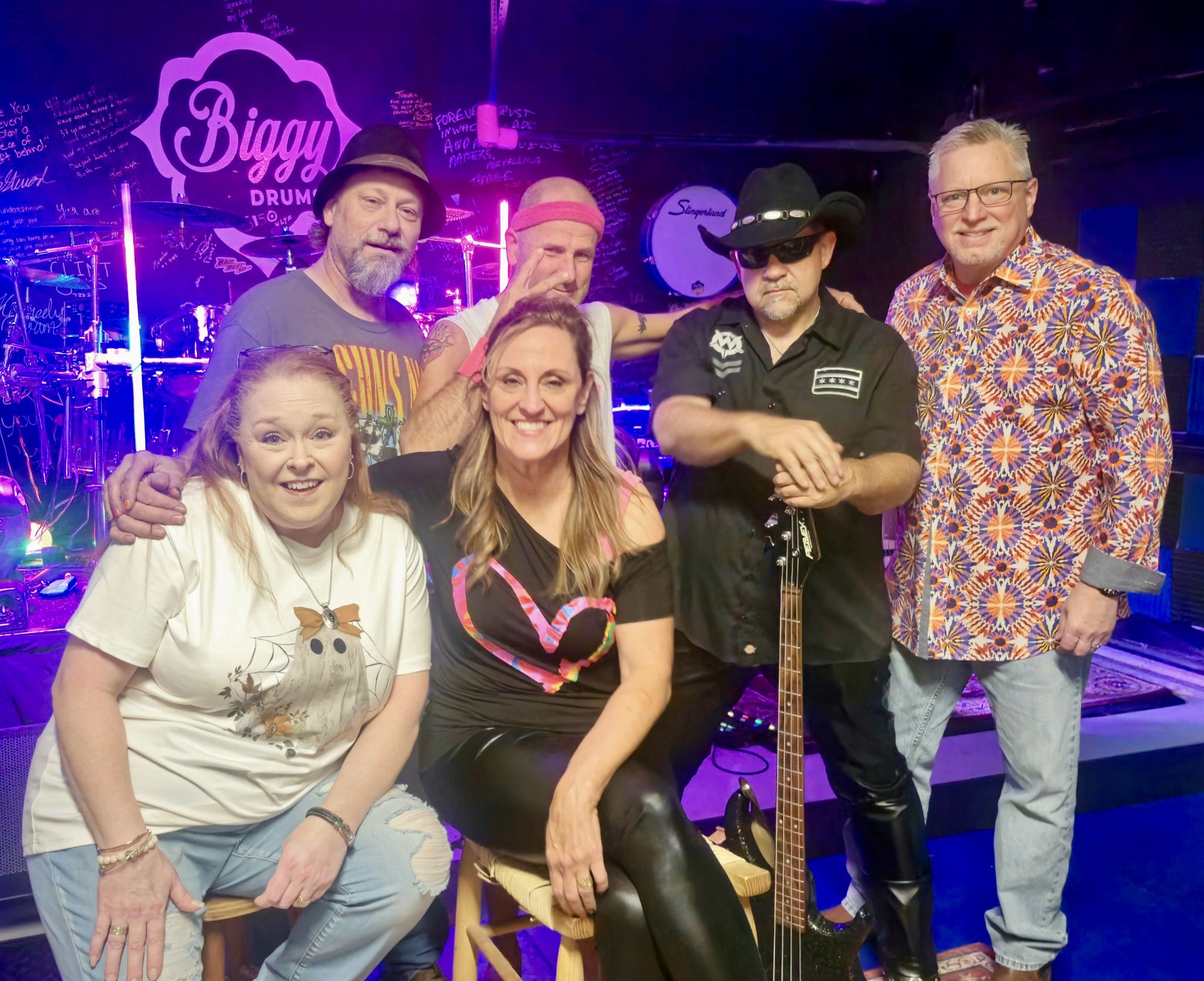 Group of six people posing on stage with musical instruments and purple neon lights behind them, including a woman seated in front, smiling.