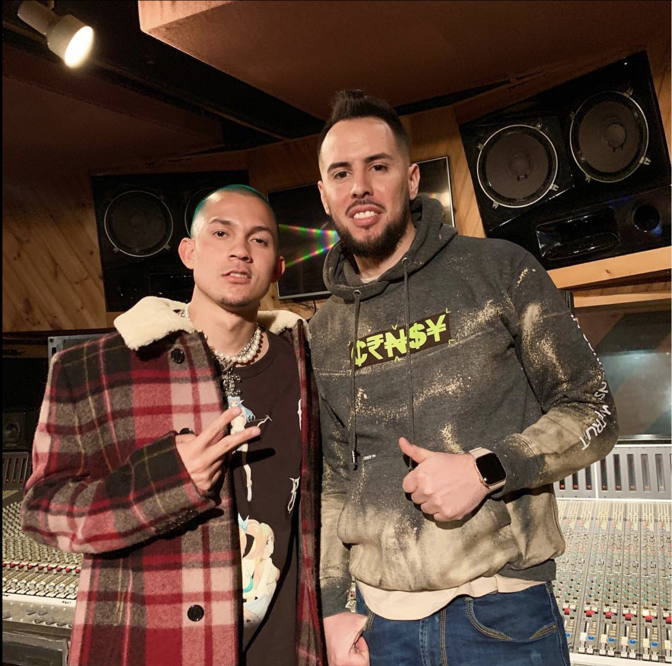 Dos hombres en un estudio de grabación, uno con abrigo de cuadros y el otro con sudadera, posando para una foto.