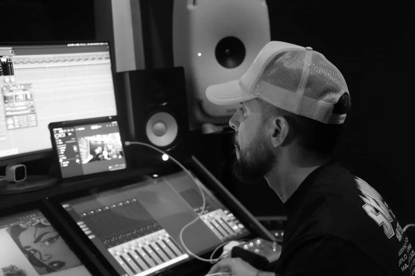 Hombre en estudio de grabación usando una computadora y equipo de audio, lleva gorra y viste ropa oscura.