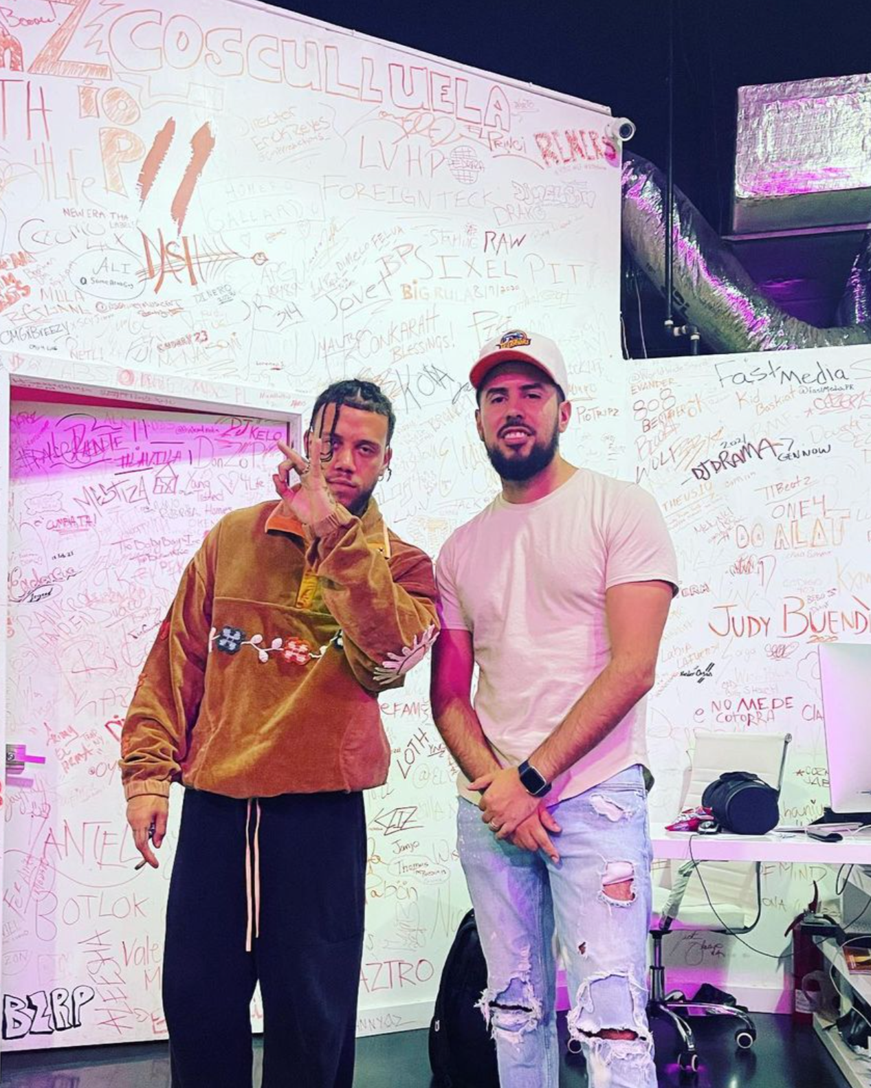 Dos hombres de pie en una pared cubierta de graffitis y escritos en colores variados, uno con suéter marrón y pantalón negro, el otro con camiseta blanca y jeans rotos, en un ambiente de estudio o sala creativa.