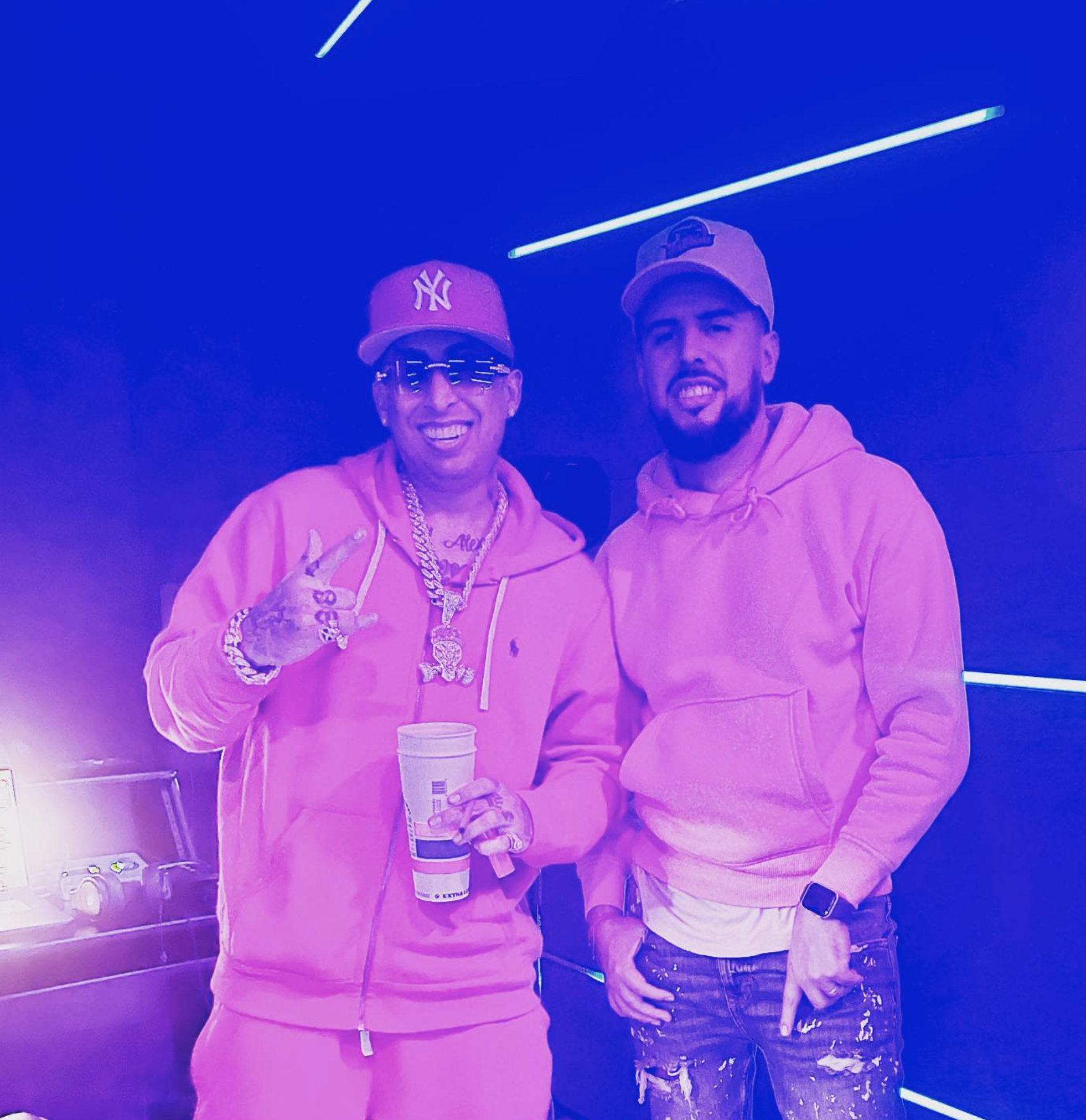 Dos hombres sonriendo en un entorno con iluminación azul, ambos llevan ropa deportiva rosa. Uno usa gafas de sol y cadenas de oro, sosteniendo un vaso de bebida.