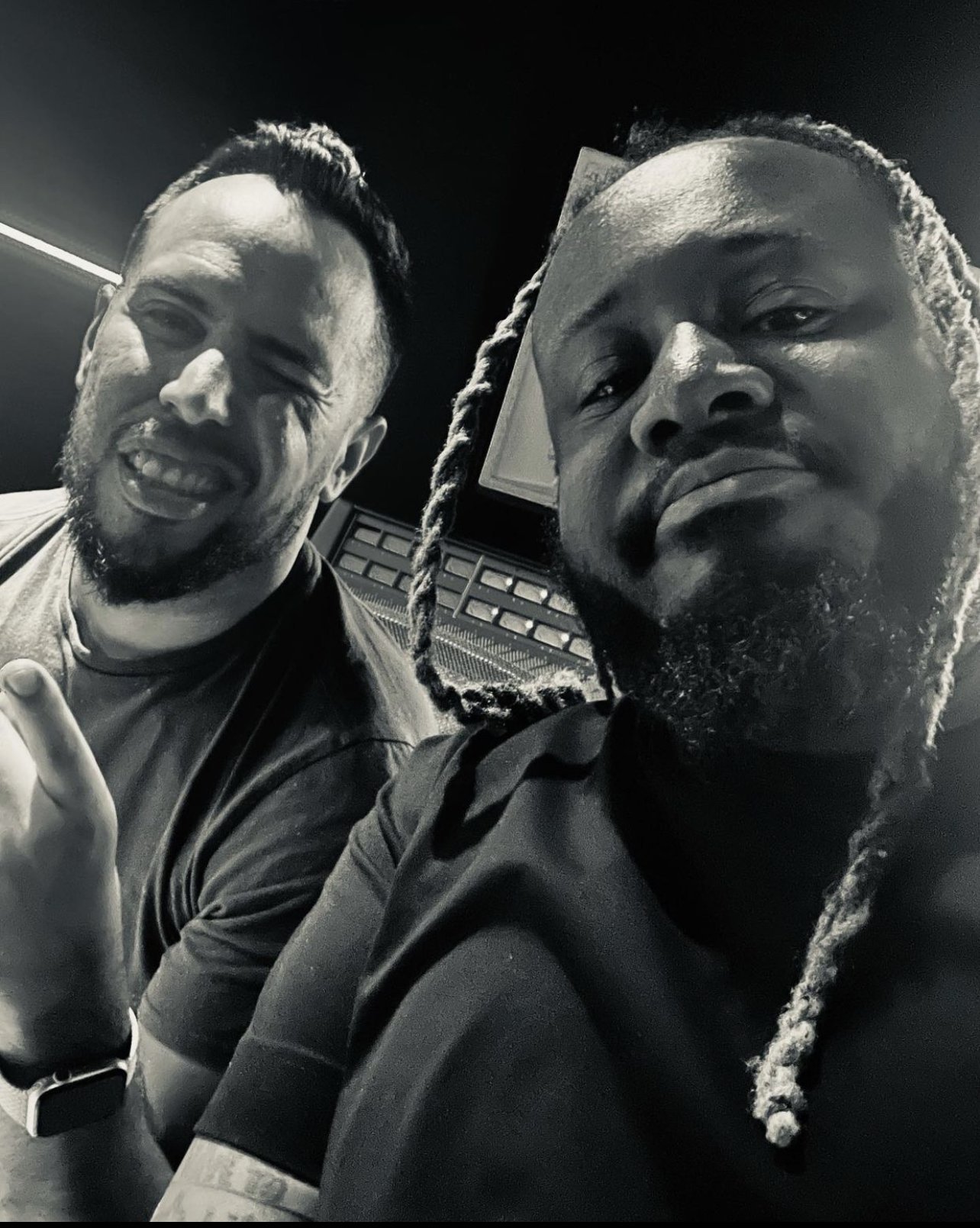 Dos hombres en un estudio de grabación posando para una selfie, uno con expresión relajada y el otro con expresión seria, ambos con barba y uno con cabello trenzado.