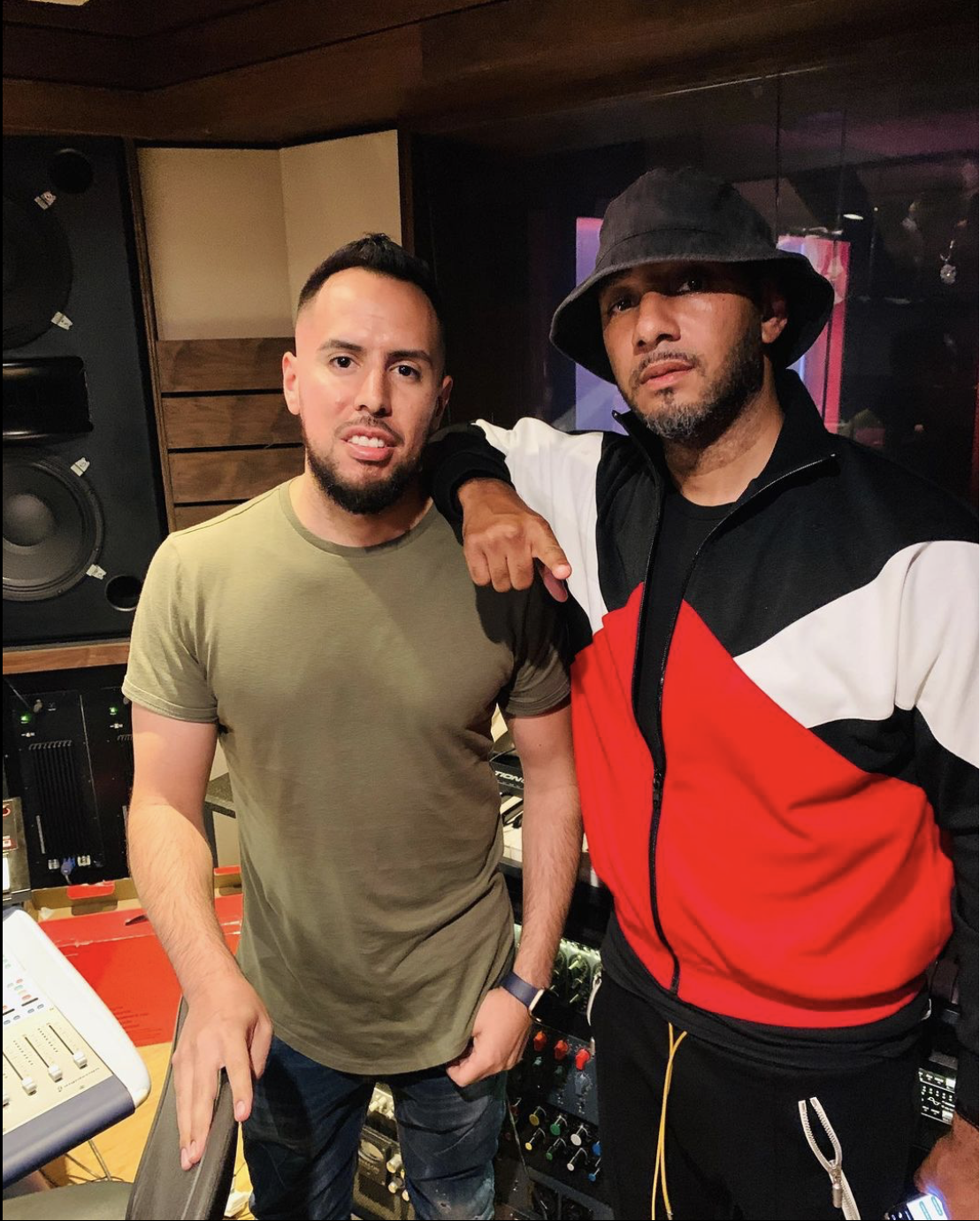 Dos hombres en un estudio de grabación, uno con camiseta verde y otro con sudadera roja y negra, el de la sudadera usa sombrero y tiene barba, ambos posando para la foto.