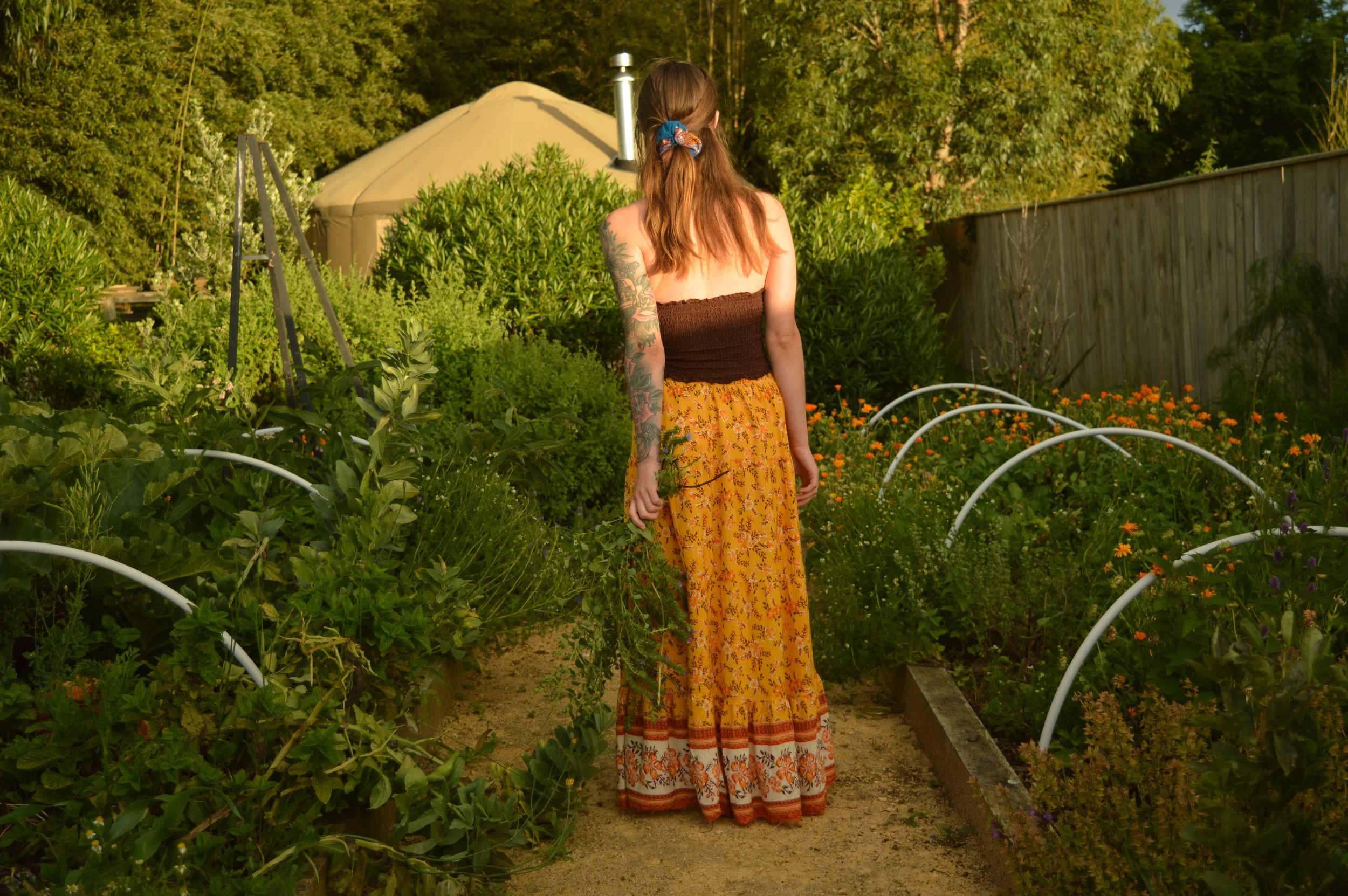 woman with tattoos on her arm from behind, standing in a garden, wearing a strapless top and a long, colorful skirt, surrounded by green plants and flowers, with a wooden fence and trees in the background
