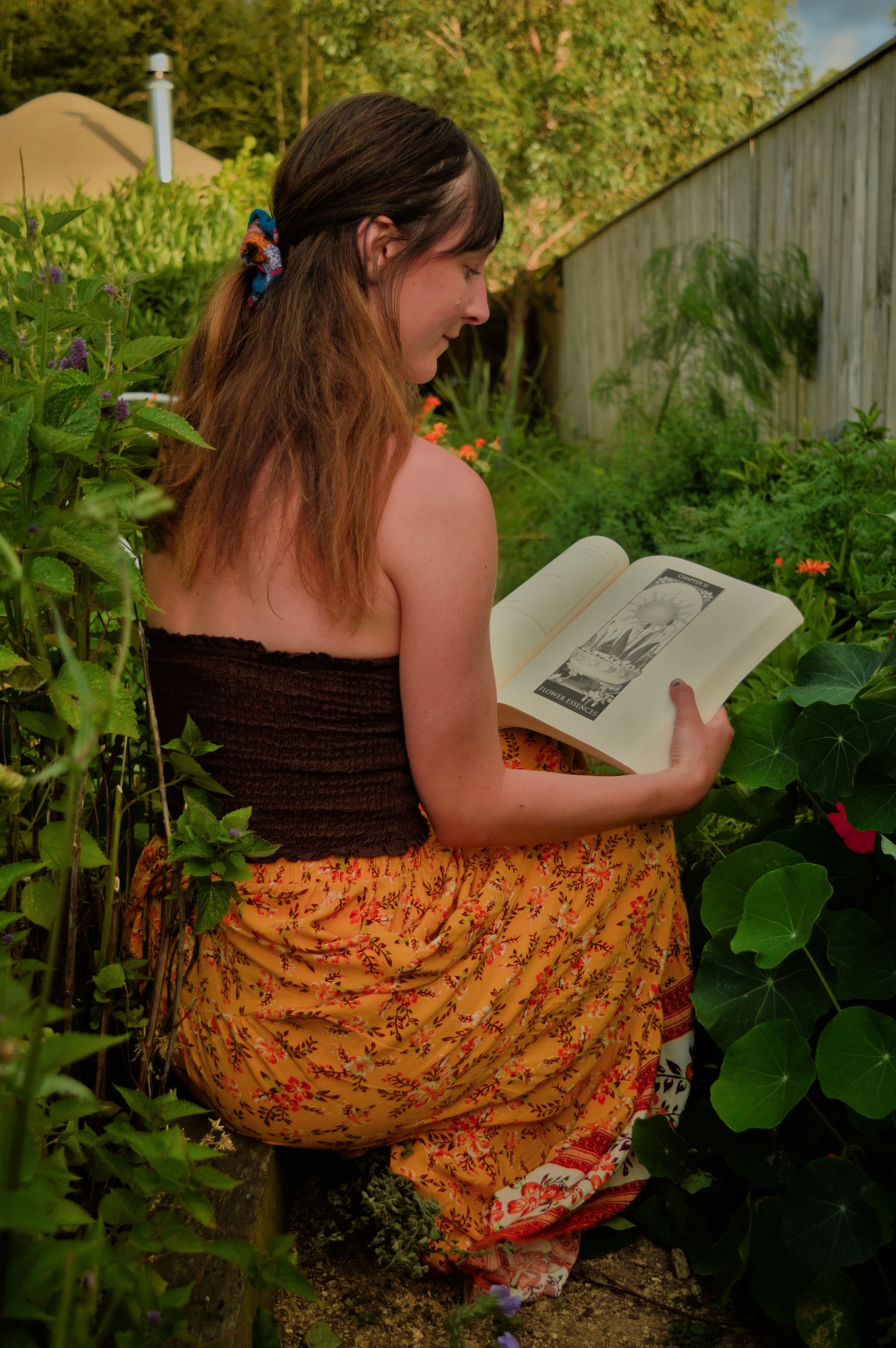 A woman sitting in a garden, reading a book about flowers.