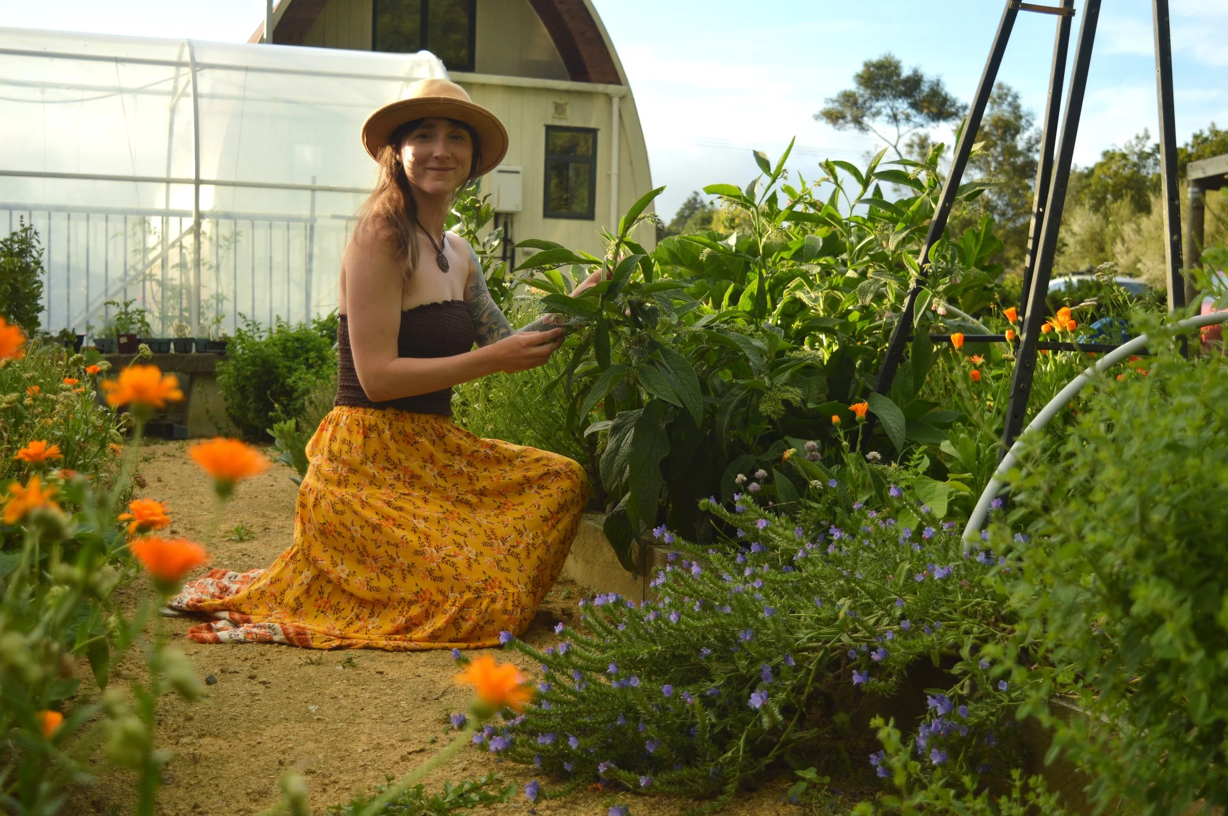 A woman kneeling in a garden surrounded by flowers and plants, wearing a wide-brimmed hat, a dark strapless top, and a yellow skirt with orange floral pattern, holding a plant.