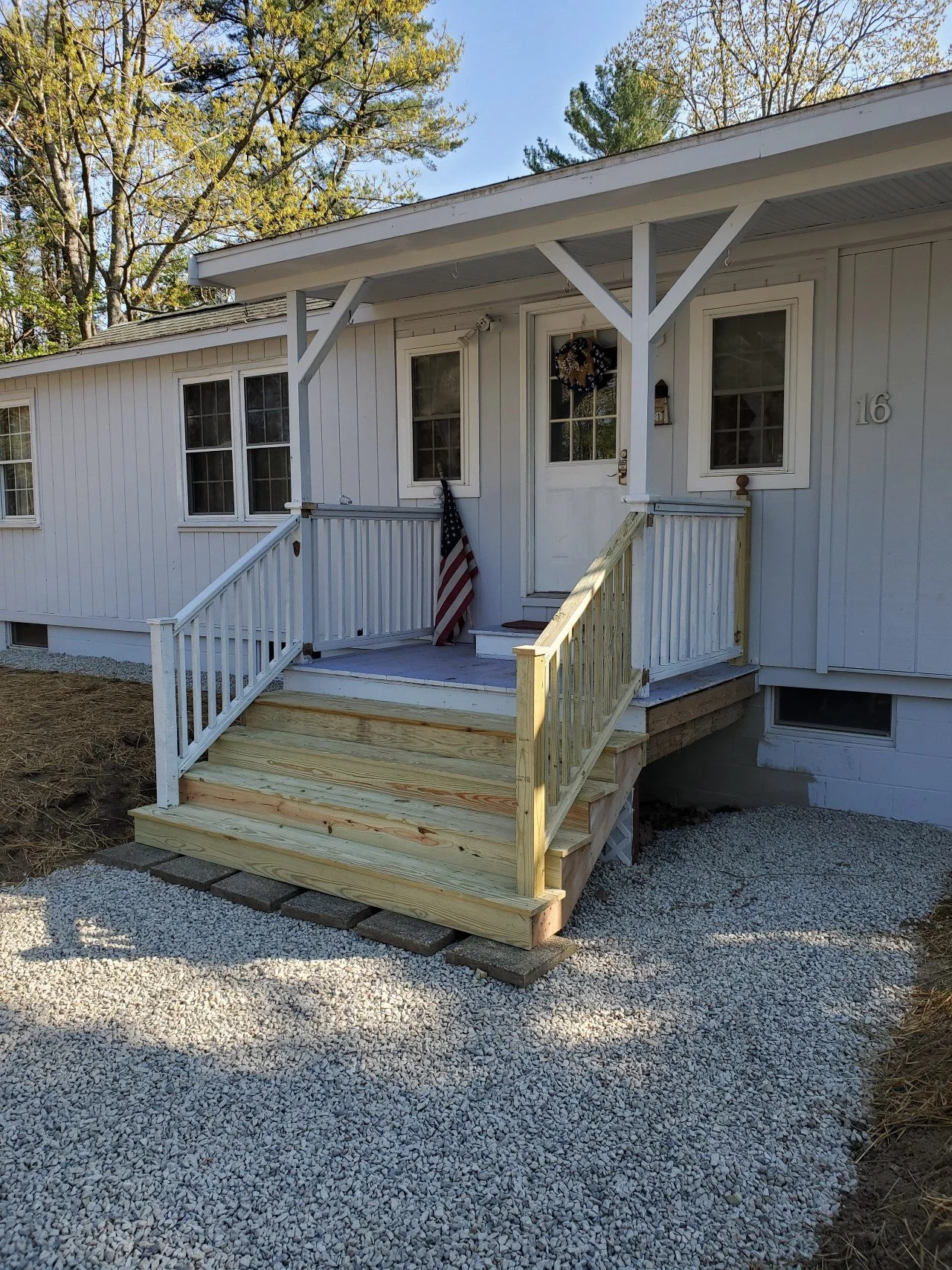 Front porch of a white house with a small set of wooden stairs, a railing, and a door decorated with a wreath. An American flag leans against the porch railing.