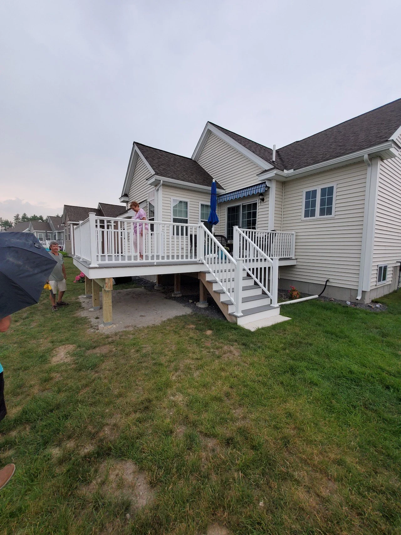 A new wooden deck with white railing attached to a house, with stairs leading down to the yard. There are people around, some standing and one woman on the deck, with a cloudy sky above.