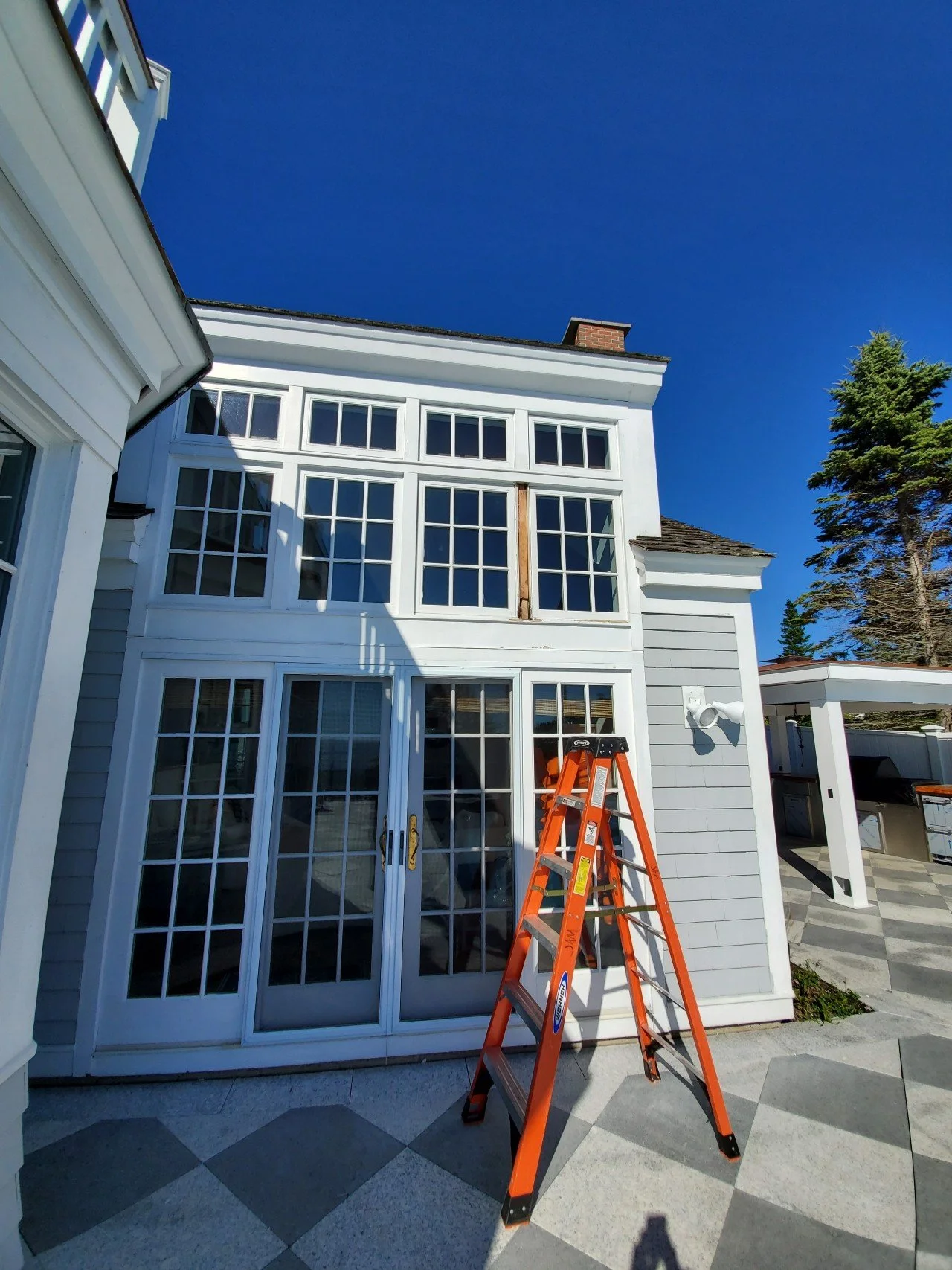 Rot repair in progress. A house with large windows, a glass double door, and an orange ladder outside under a clear blue sky.