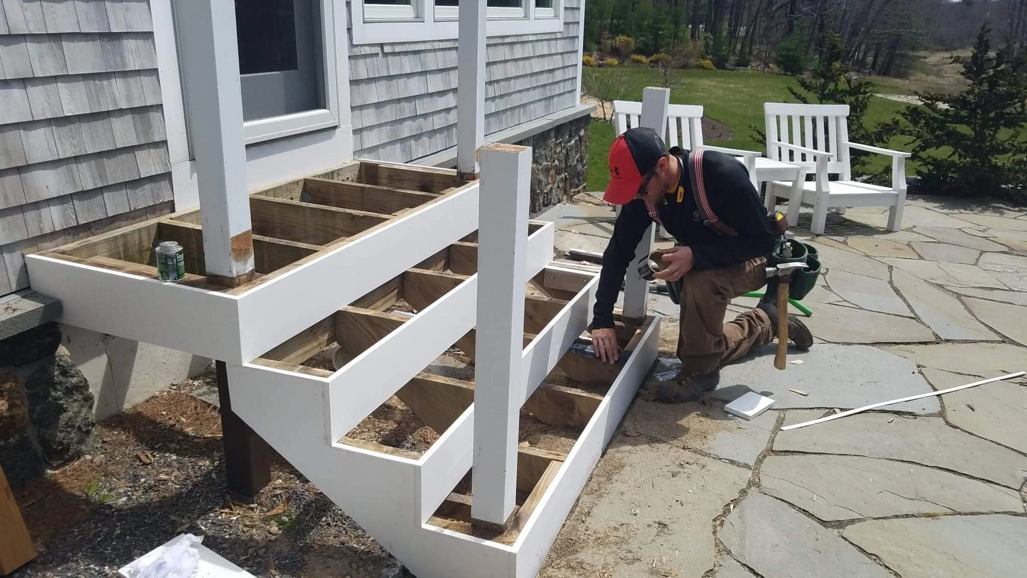 A person working on building a white wooden staircase outside a house, with some chairs and a garden in the background.