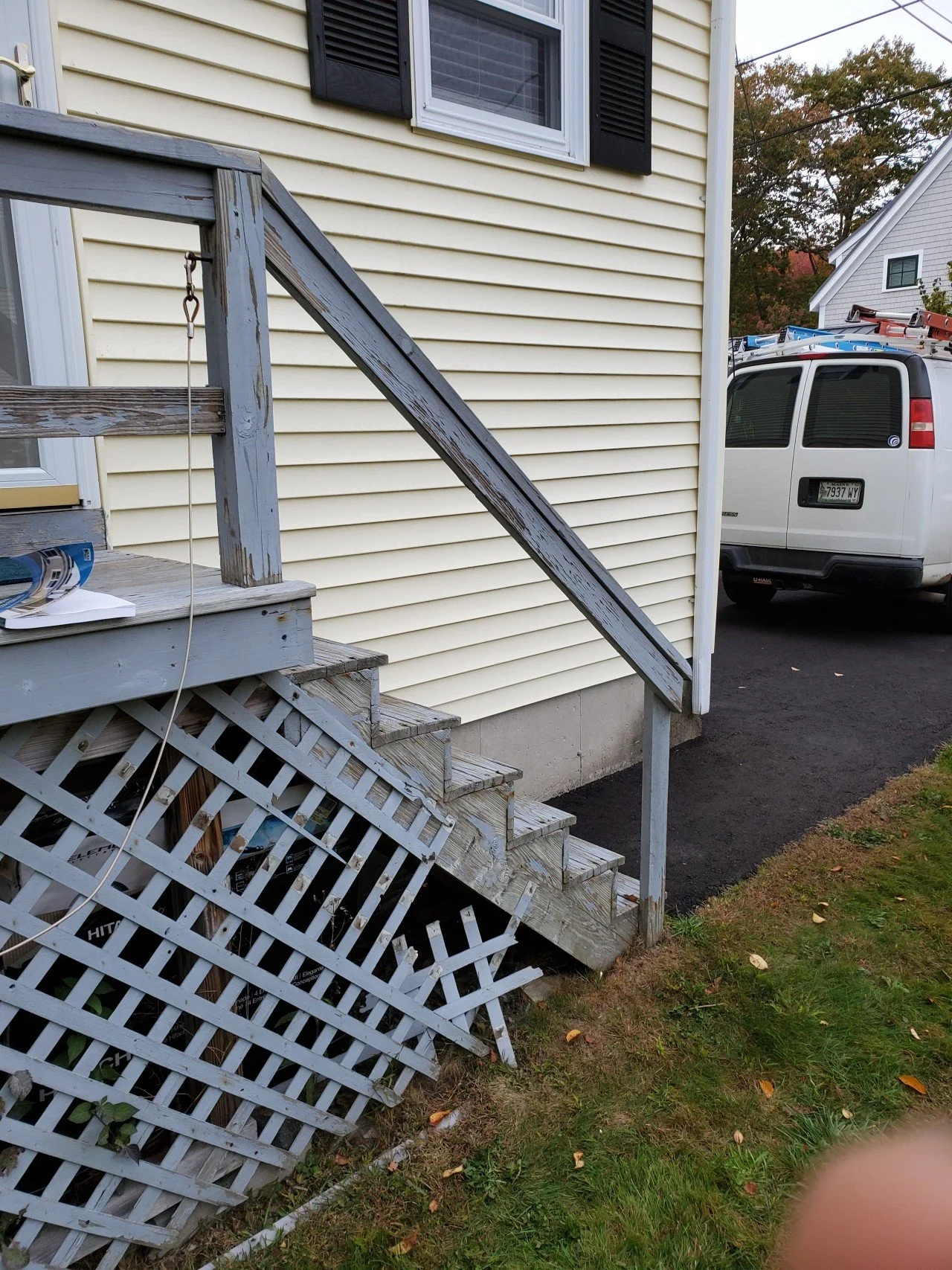 Backyard stairs of a house with damaged lattice, worn gray wood steps, and a white house with black window shutters. A white van is parked nearby with ladders on top, and trees are visible in the background. Before porch was replaced.