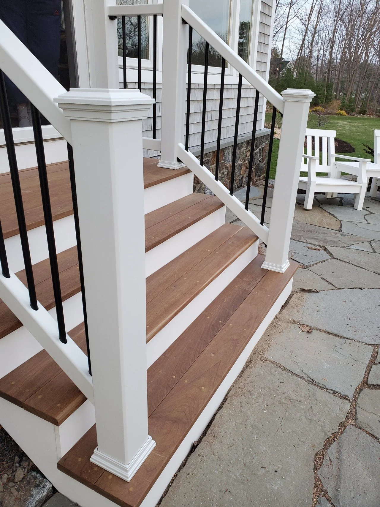 After photo of mahogany porch staircase with white trim, black metal railings, and a stone foundation, leading to a patio with outdoor chairs and trees in the background.