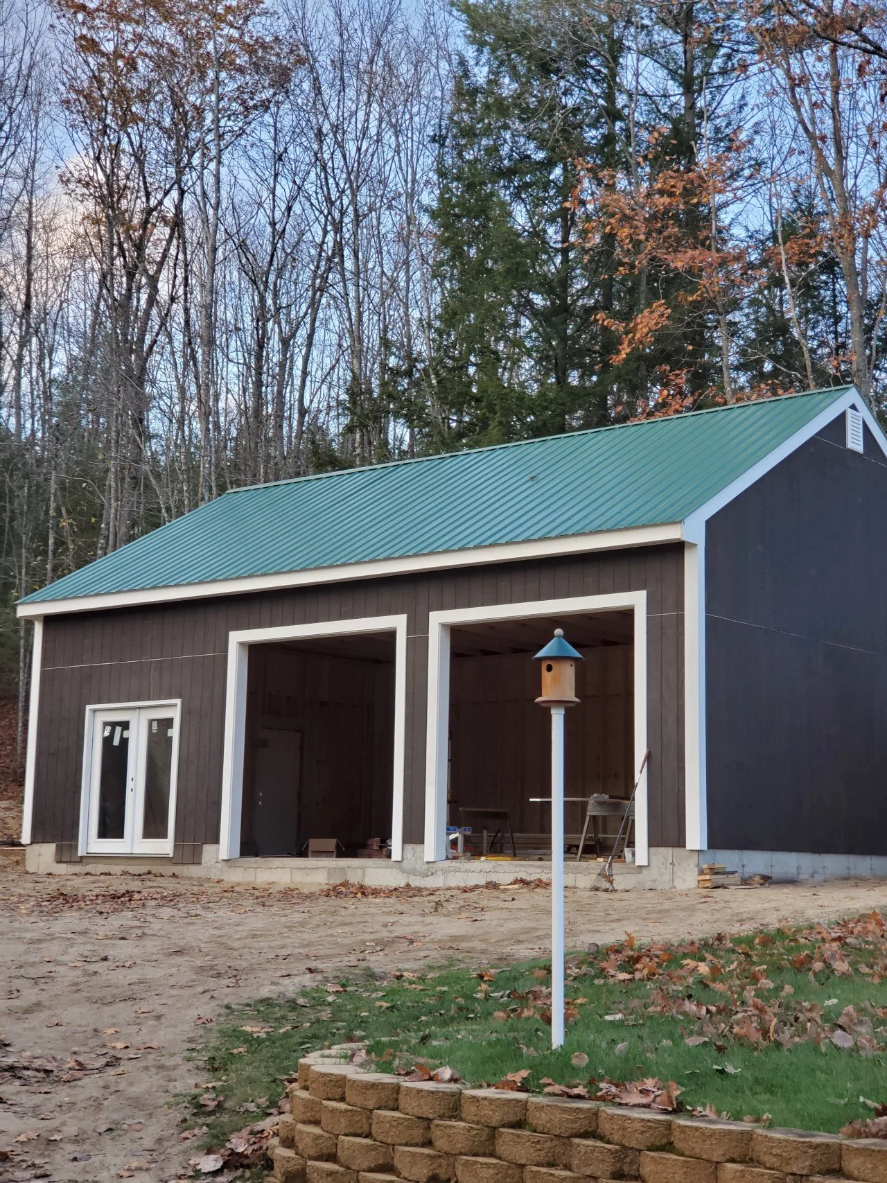 A finished three car garage with a green metal roof, black siding, and large open garage spaces. There is a birdhouse on a post in front of the house, surrounded by a brick-bordered flower bed with fallen leaves. 