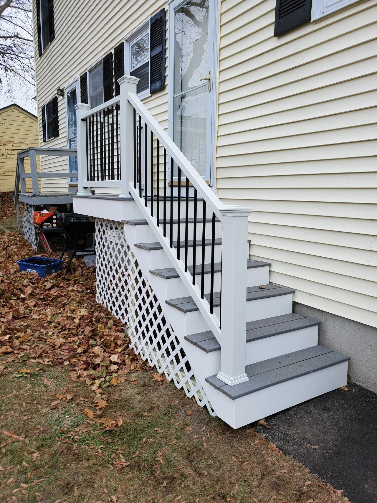 New front porch stairs with white railing and gray steps leading to a glass door, adjacent to light yellow house with black shutters.