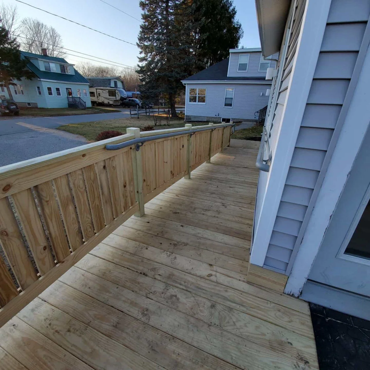 Newly constructed wooden handicap ramp attached to a local business, with a partial view of the business's exterior siding, overlooking a neighborhood street with houses, trees, and parked vehicles.