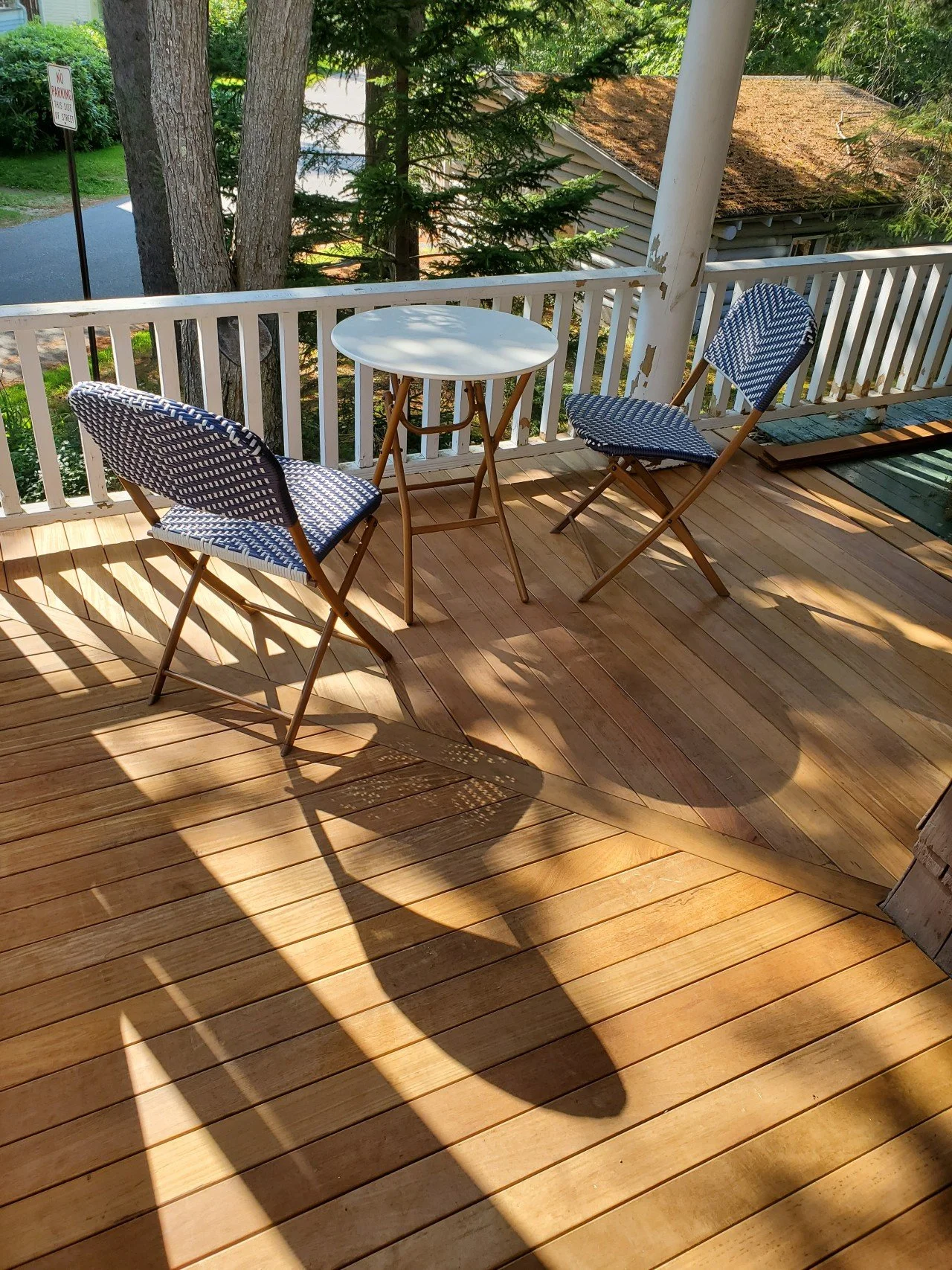 Mahogany deck with a small white round table and two foldable chairs with blue and white patterned cushions, surrounded by a white railing, with trees and a house in the background, and shadows cast on the floor.