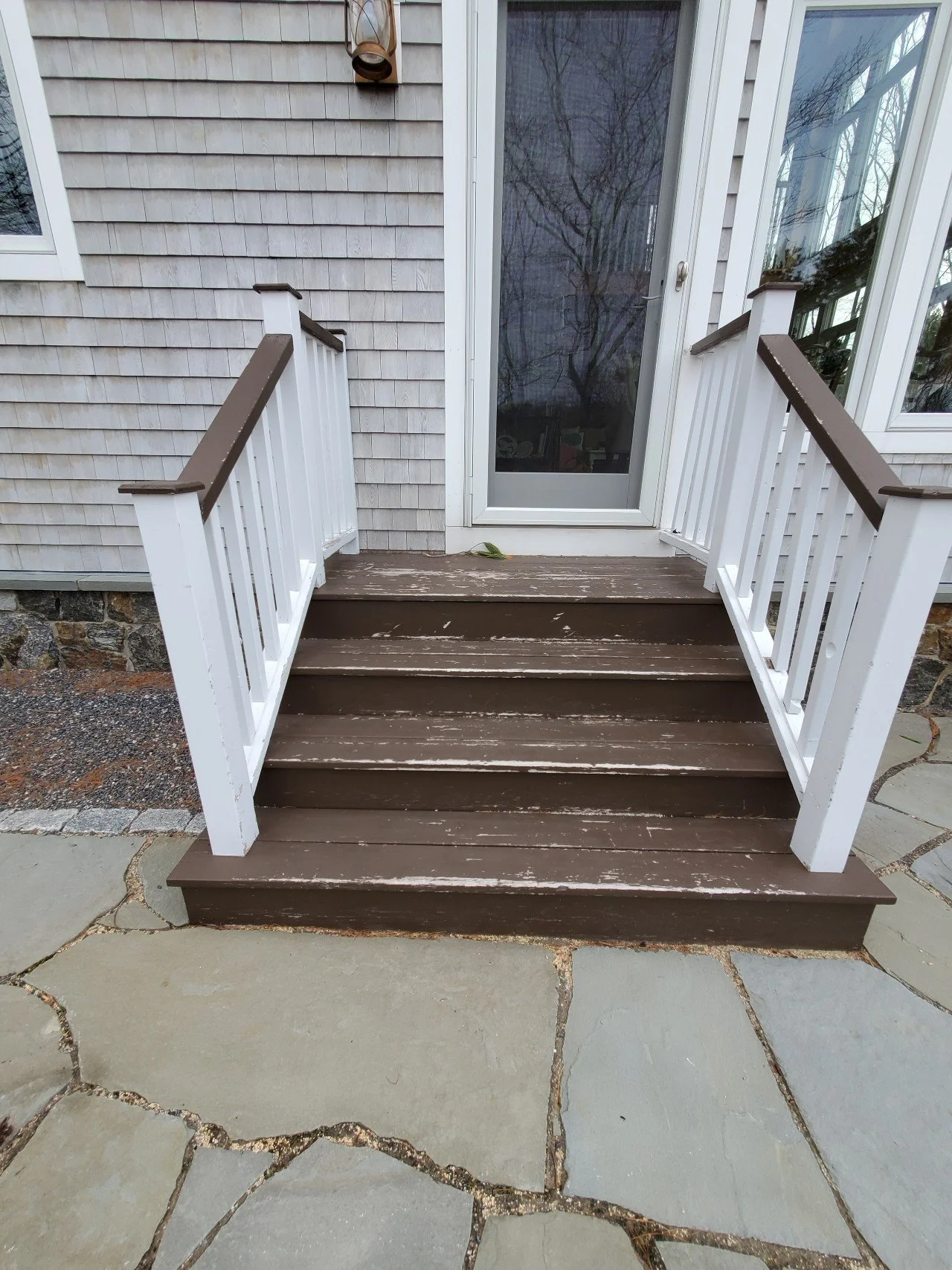 Before view of a small wooden porch with brown steps and white railing, leading to a glass door on the house, with a stone and concrete pathway in front.