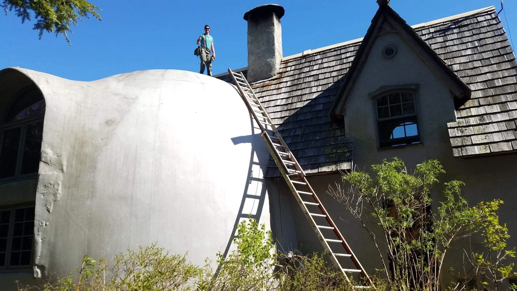 A man standing on the roof of a dome house with a ladder leaning against a rounded, white addition. Roof repair in progress