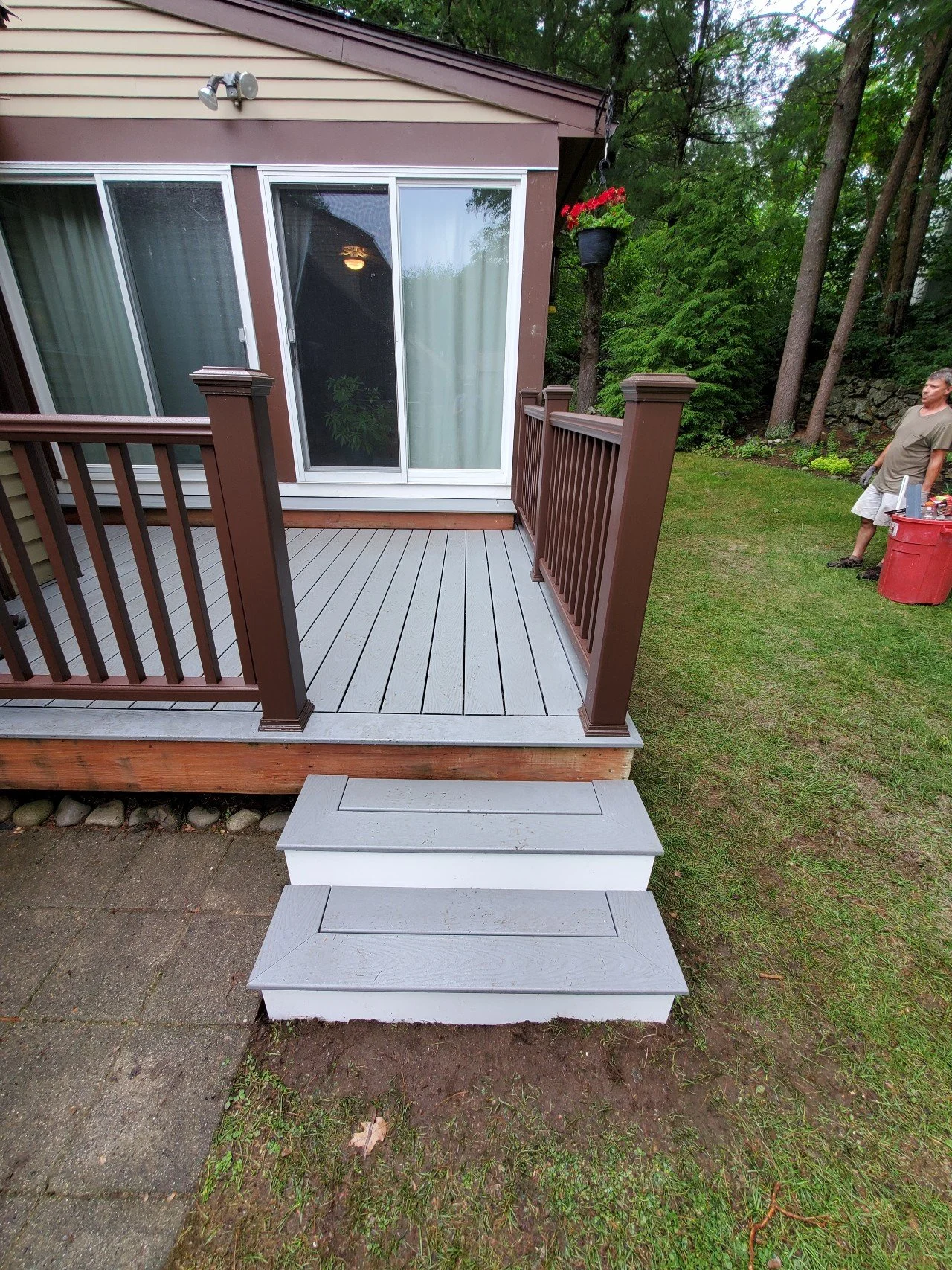 A small outdoor deck attached to a house with sliding glass doors and brown railings. There are two small stairs leading up to the deck. To the right, a man is standing near a red trash bin on a grassy yard with trees in the background.