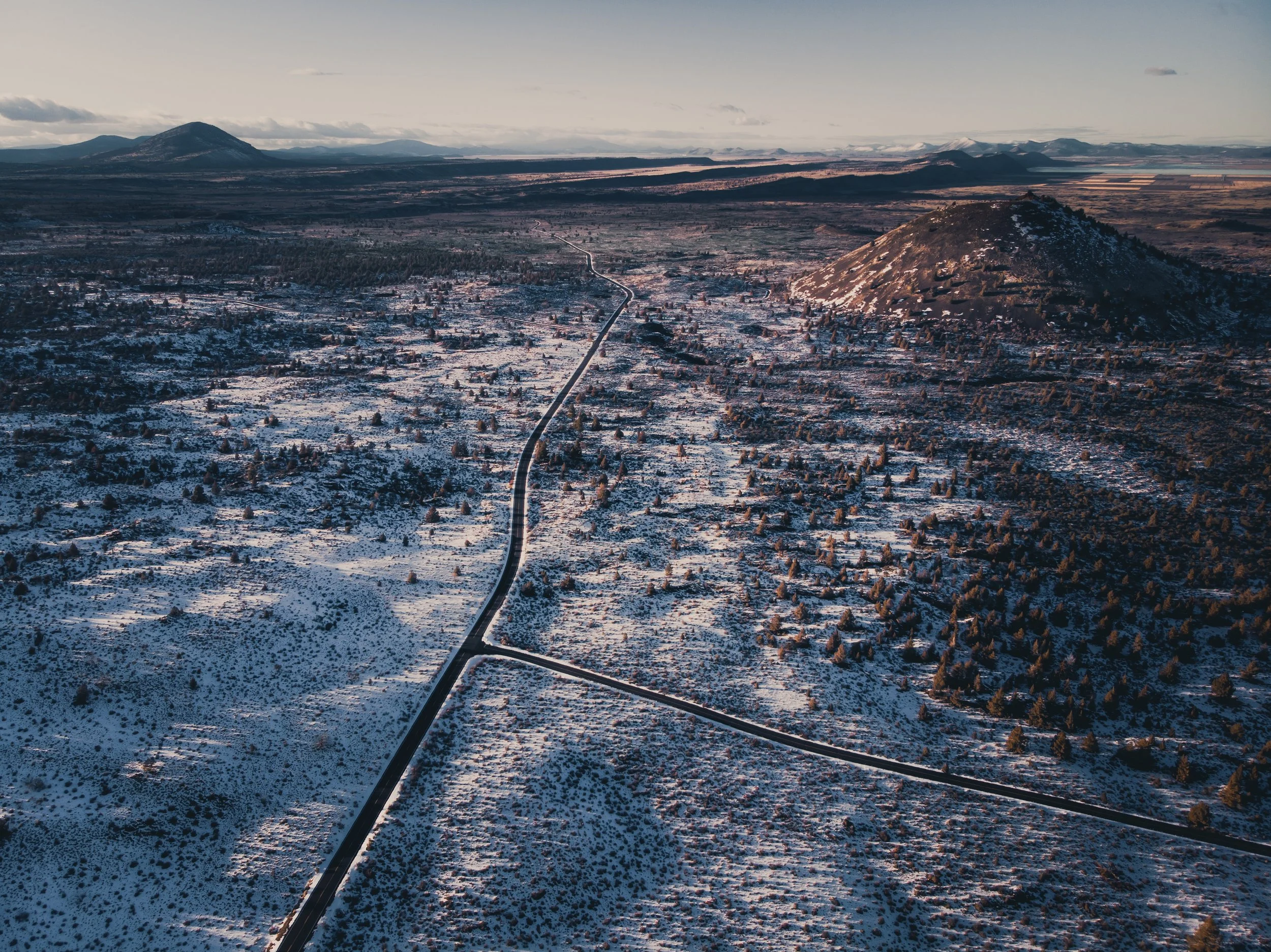 Aerial view of a snow-covered landscape with roads, scattered trees, and a hill, with mountains in the background under a cloudy sky.