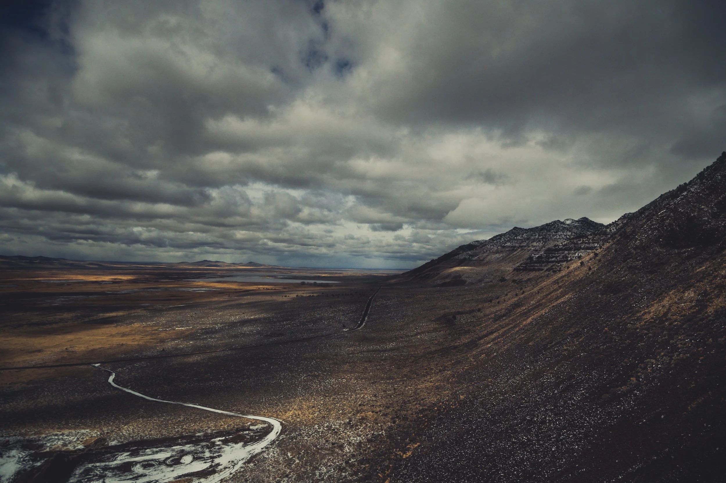 A vast, open landscape with dark, cloudy skies overhead. There are mountains on the right side and a winding dirt road in the foreground leading toward the horizon.