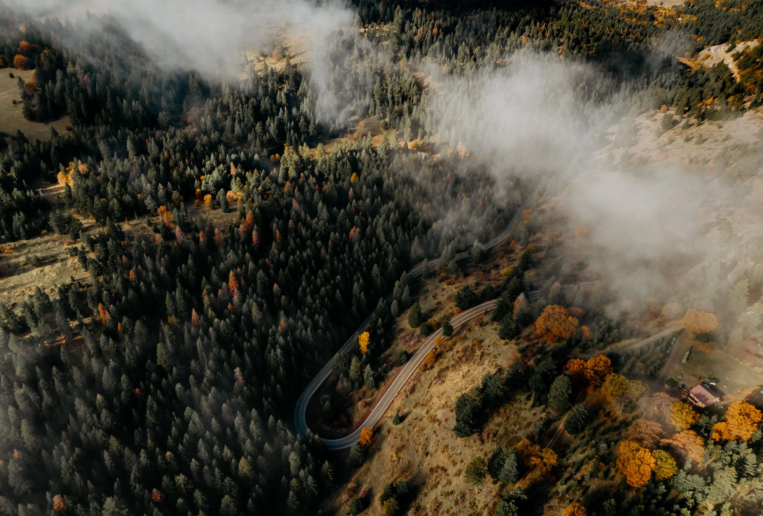 Aerial view of a winding mountain road through a dense forest with patches of orange and yellow autumn trees, some fog or smoke partially obscuring the scenery.