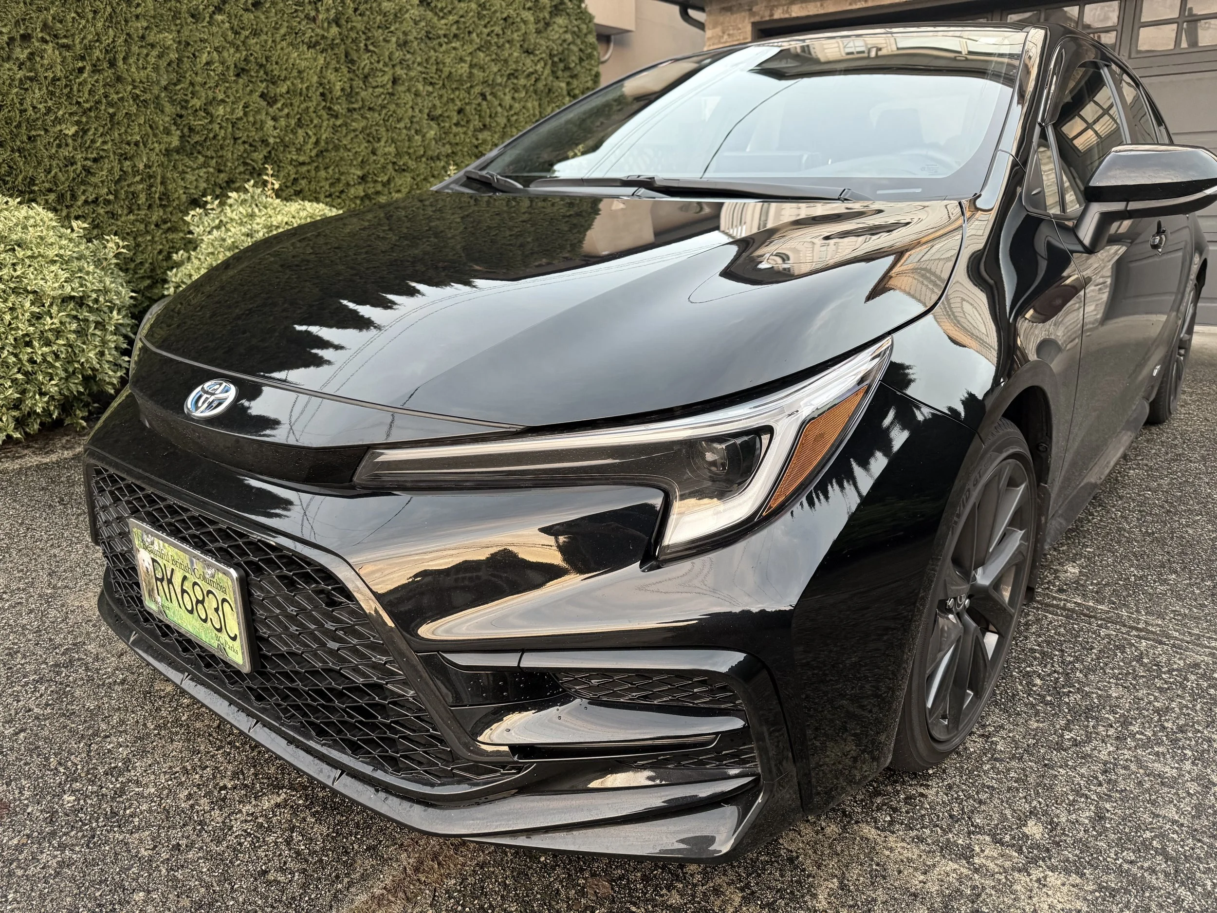 A black Toyota car parked on a paved driveway next to green bushes with a beige building reflected on the windshield and hood.