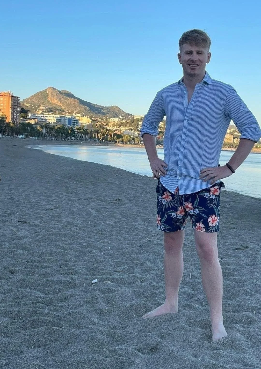 A man standing on a sandy beach with a mountain and city skyline in the background, wearing a blue striped shirt and floral swim trunks, smiling with hands on hips.