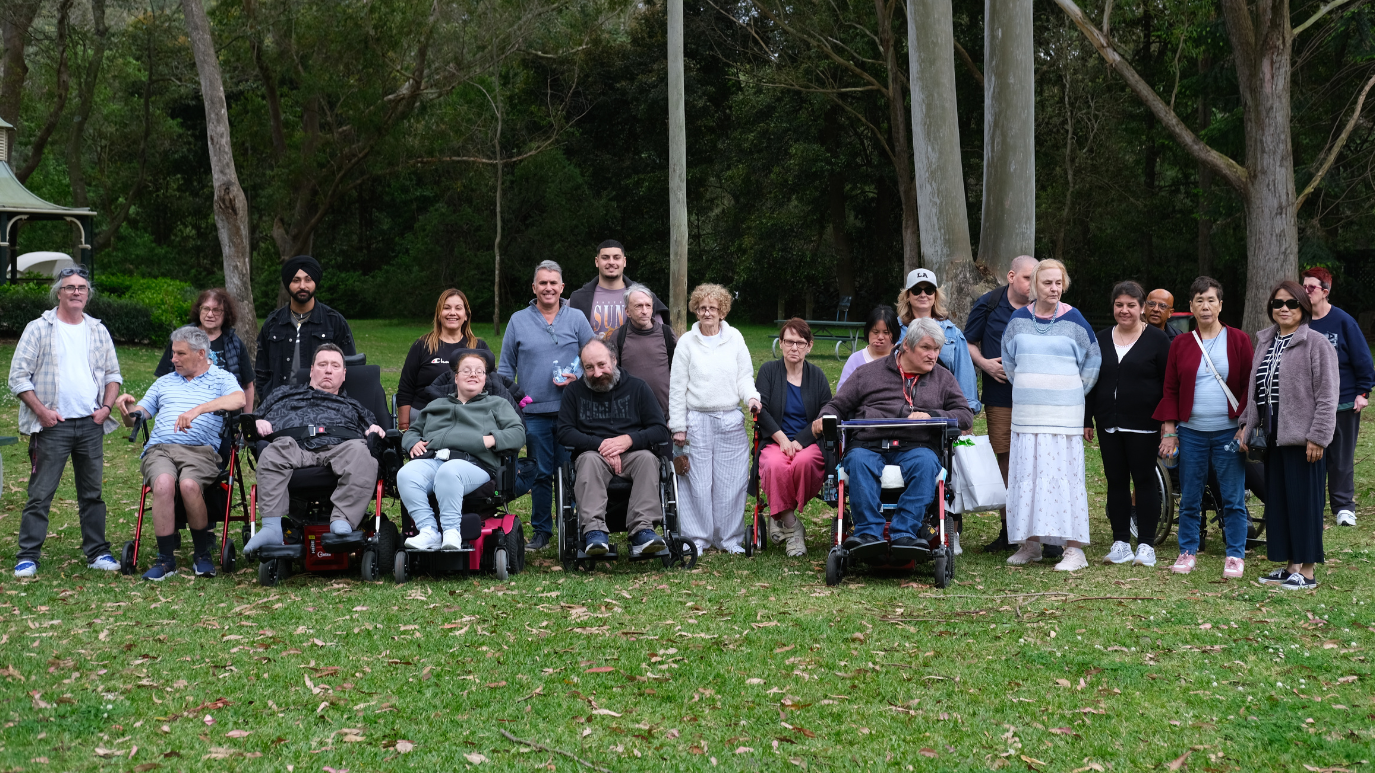 Group of diverse adults, some in wheelchairs, gathered outdoors in a park with green grass and tall trees.
