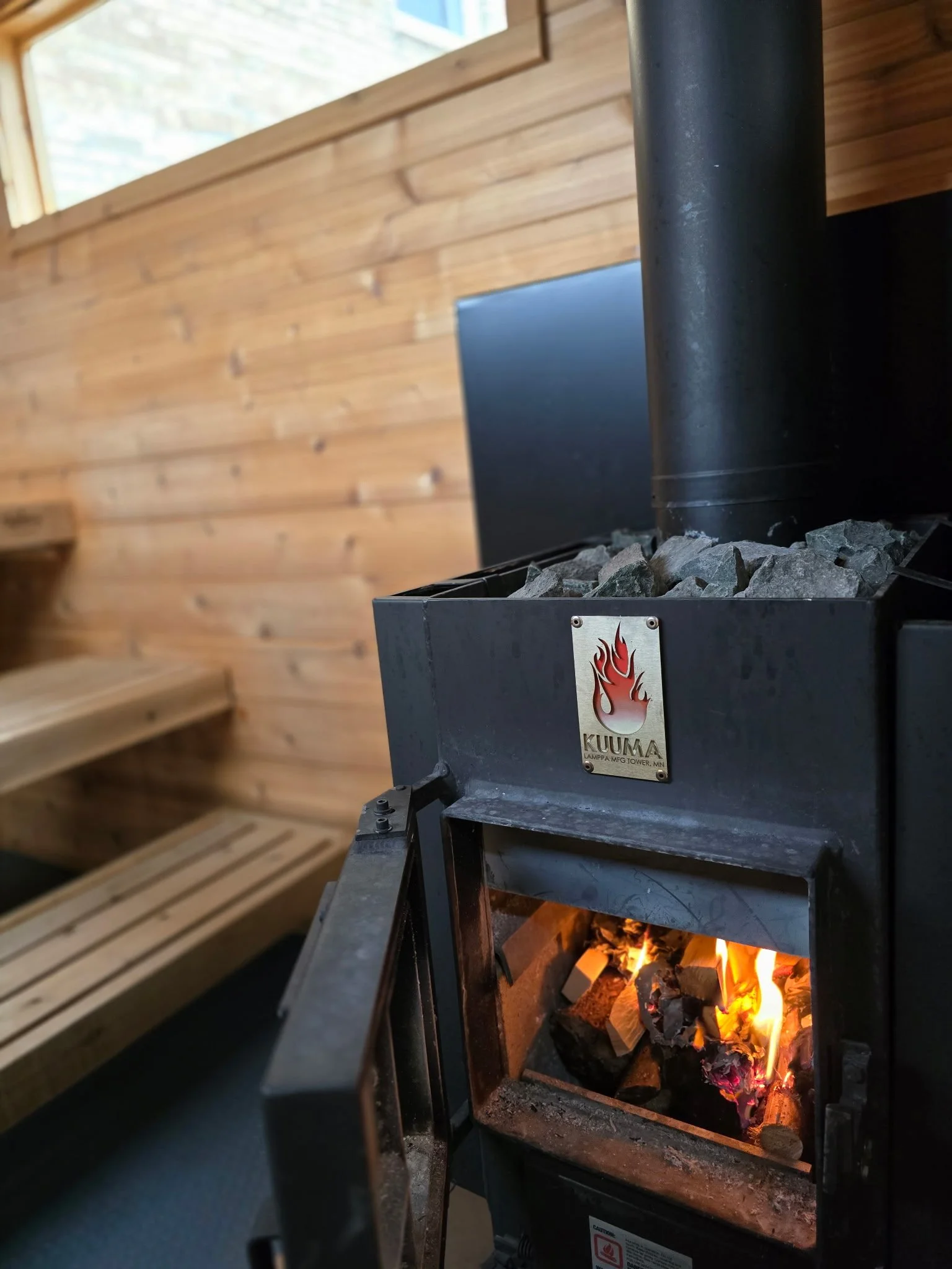 A close-up of a black Kumma wood-burning stove with a fire inside, set against a wooden interior wall with wooden stairs visible on the left.