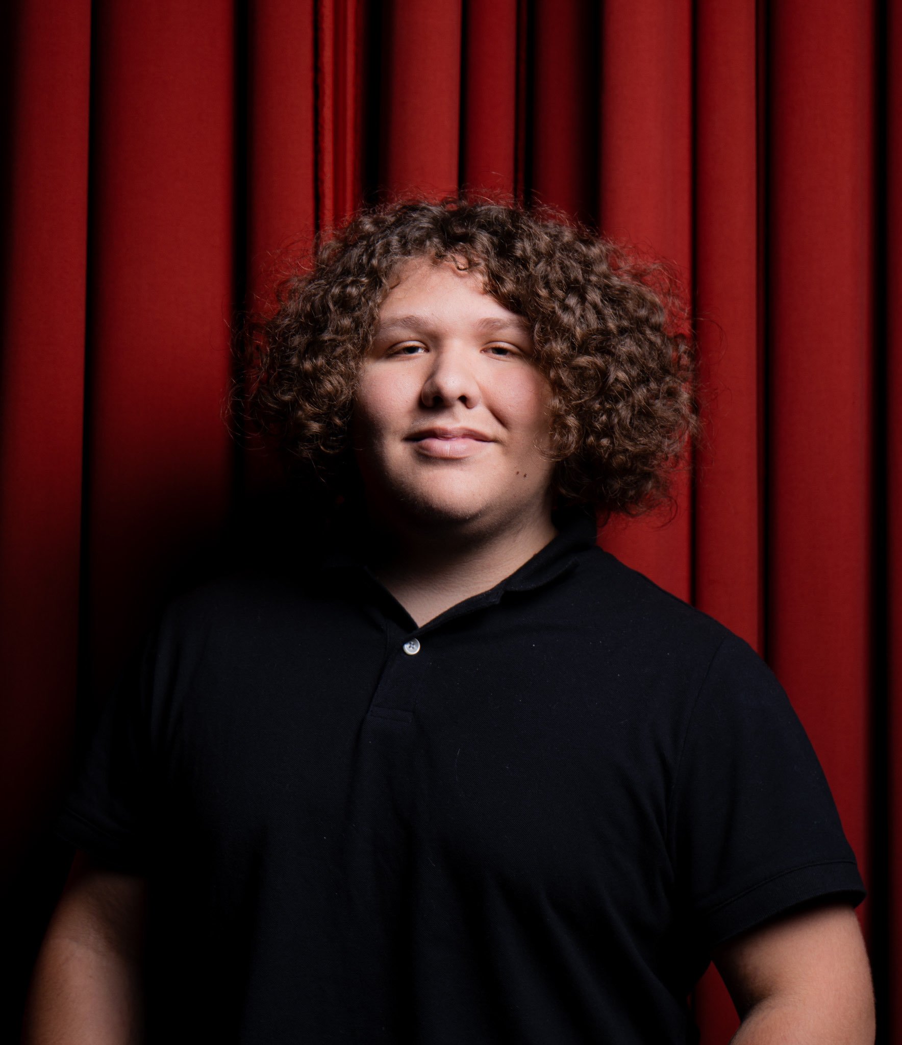 Portrait of a young man with curly hair, black shirt, standing in front of red curtain.