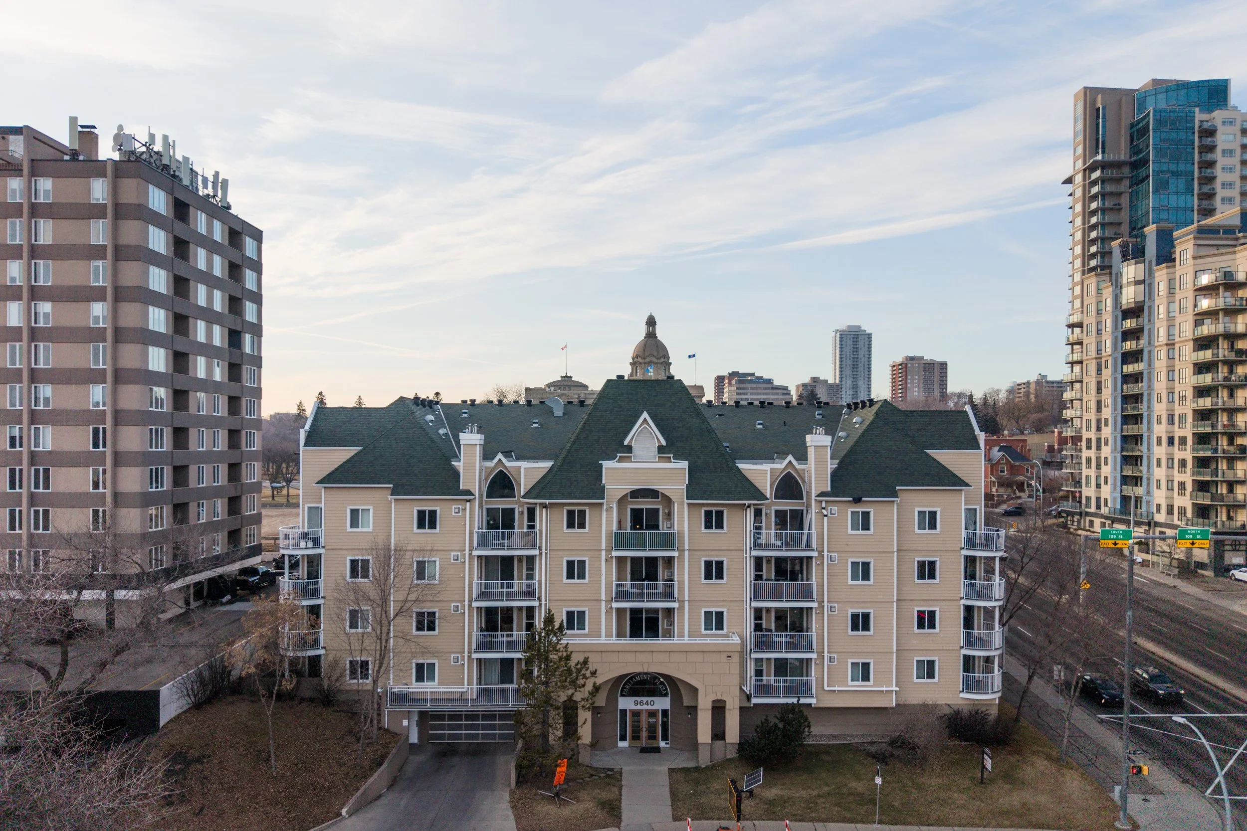 A large beige and white multi-story apartment building with a green roof, surrounded by taller modern buildings in an urban setting under a partly cloudy sky.