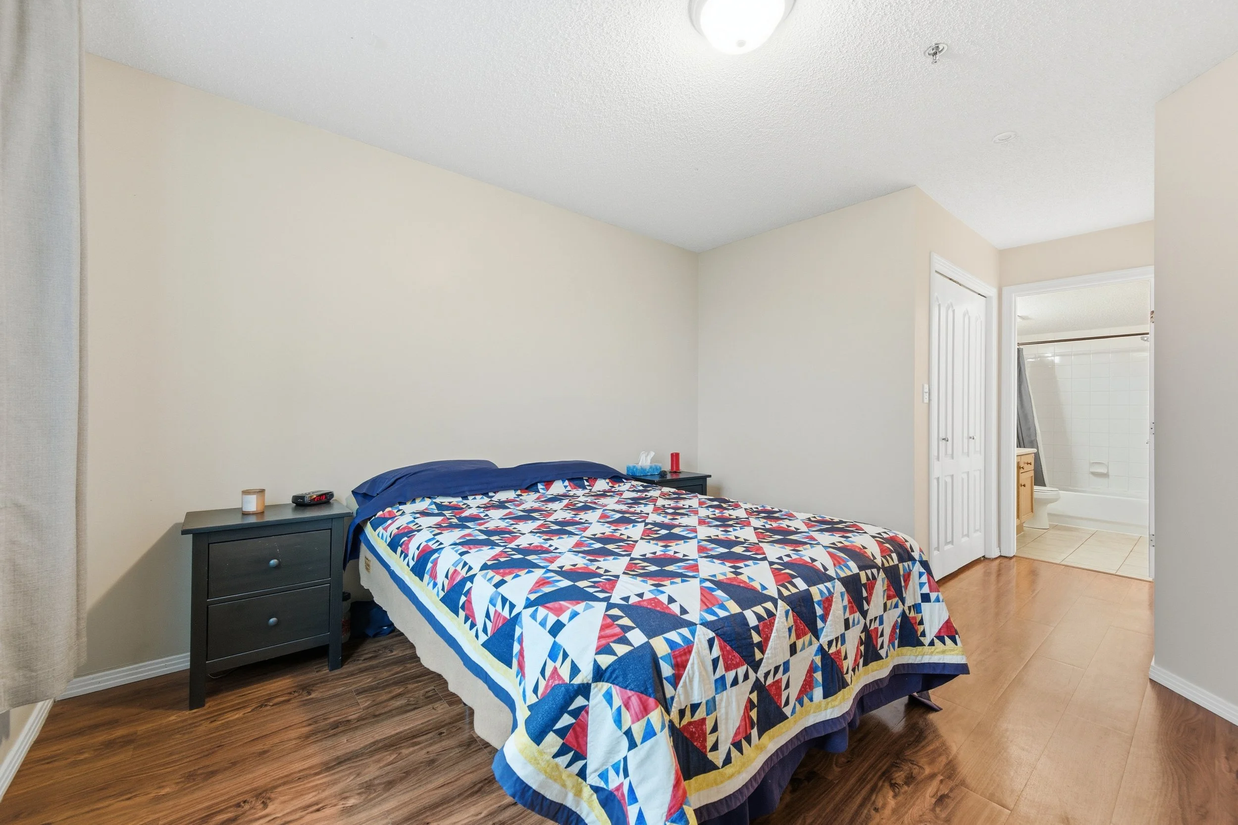 A bedroom with a bed covered in a colorful quilt, two black nightstands, and a doorway leading to a bathroom with a toilet and shower.