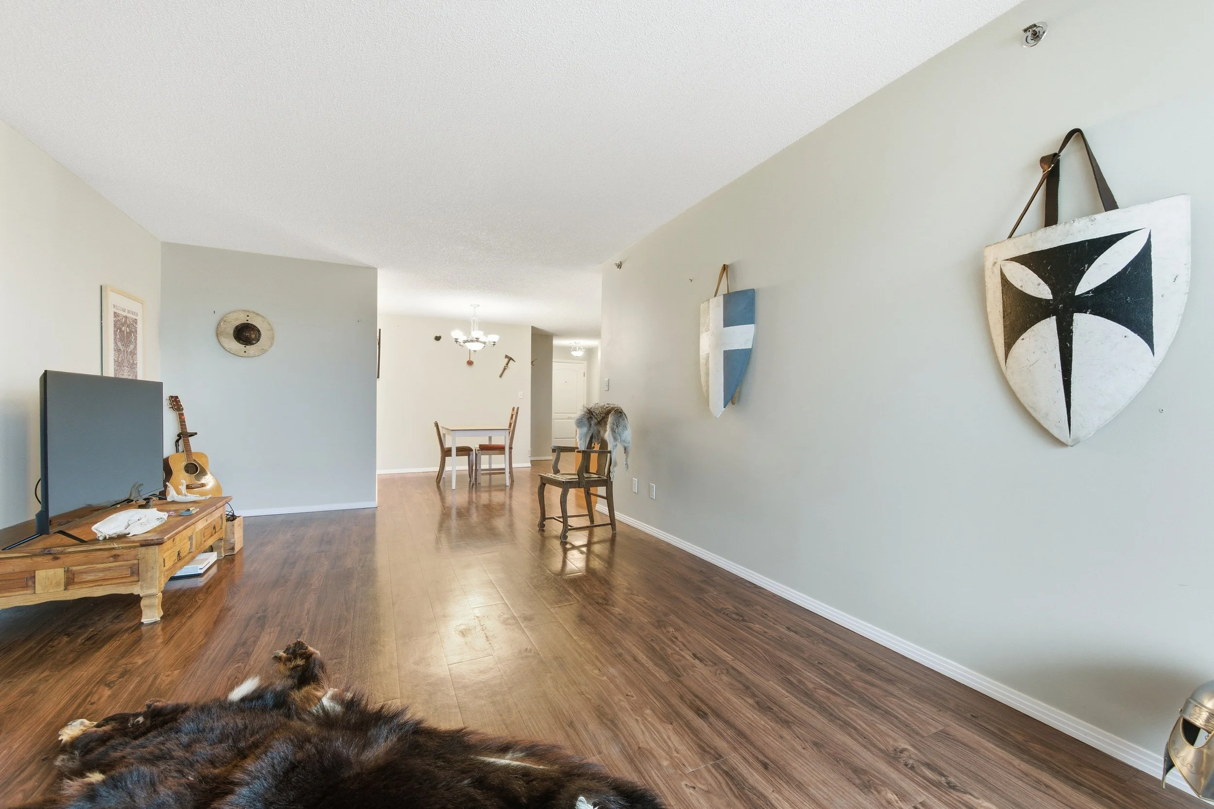 Spacious living room with hardwood floors, decorated with medieval shields on the wall, a dog lying on the floor, and a dining area in the background.