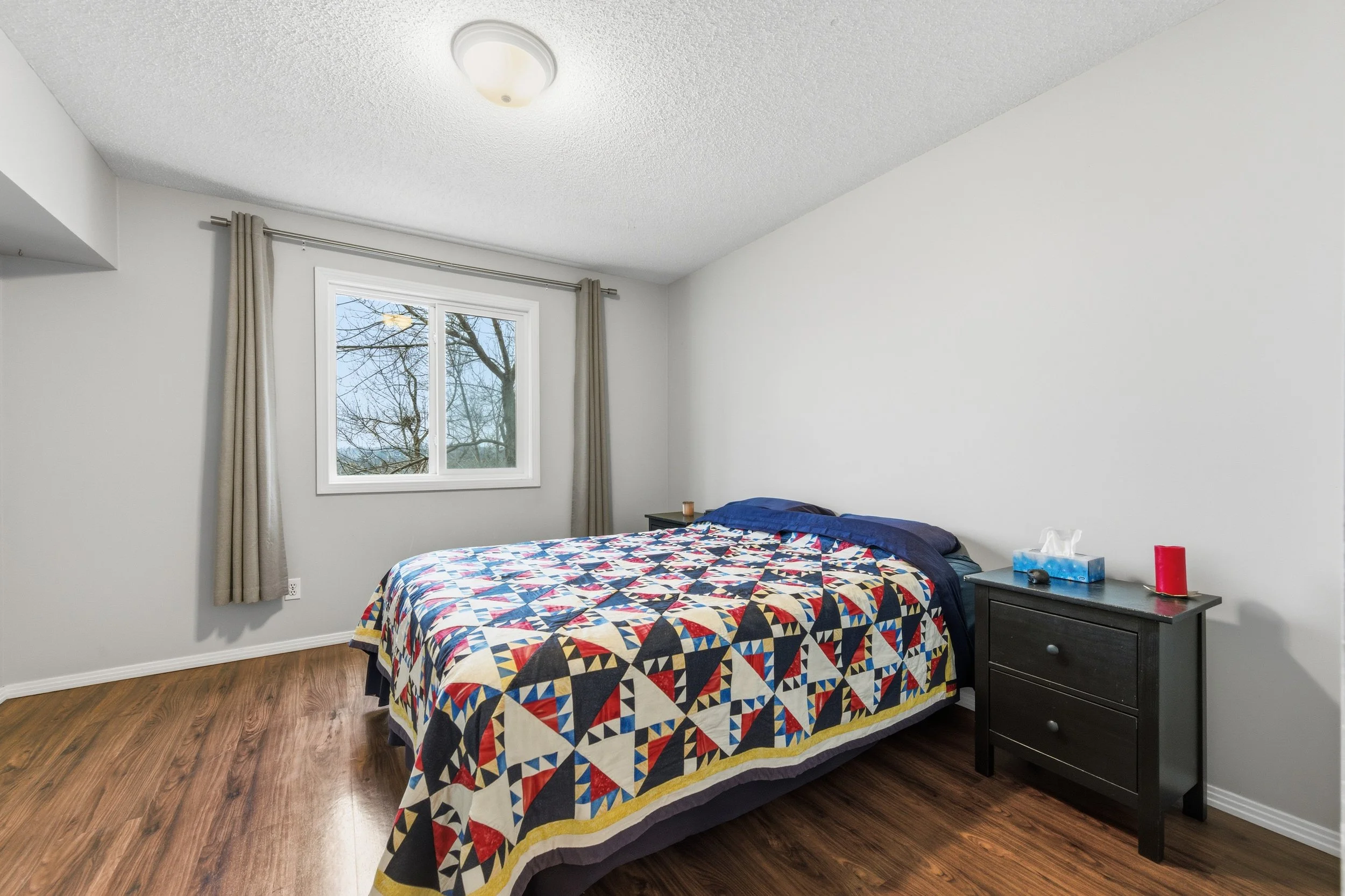 Bedroom with a bed covered by a colorful quilt, a window with beige curtains, a black nightstand holding tissues, a candle, and a red container, and wooden flooring.
