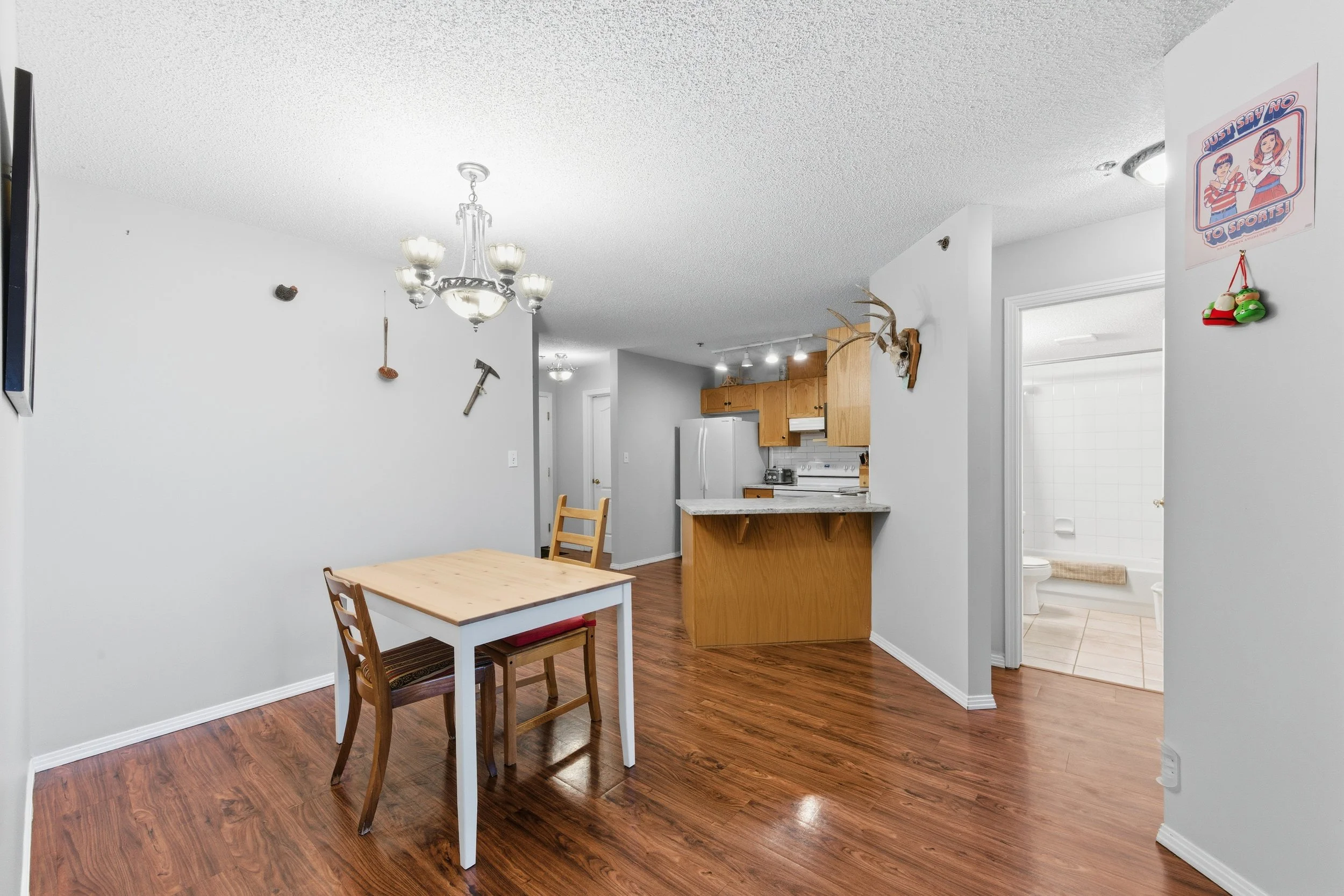 Dining area with a small wooden table and two chairs, near an open kitchen with wood cabinets and white appliances, adjacent to a bathroom with white tiled walls and a toilet.