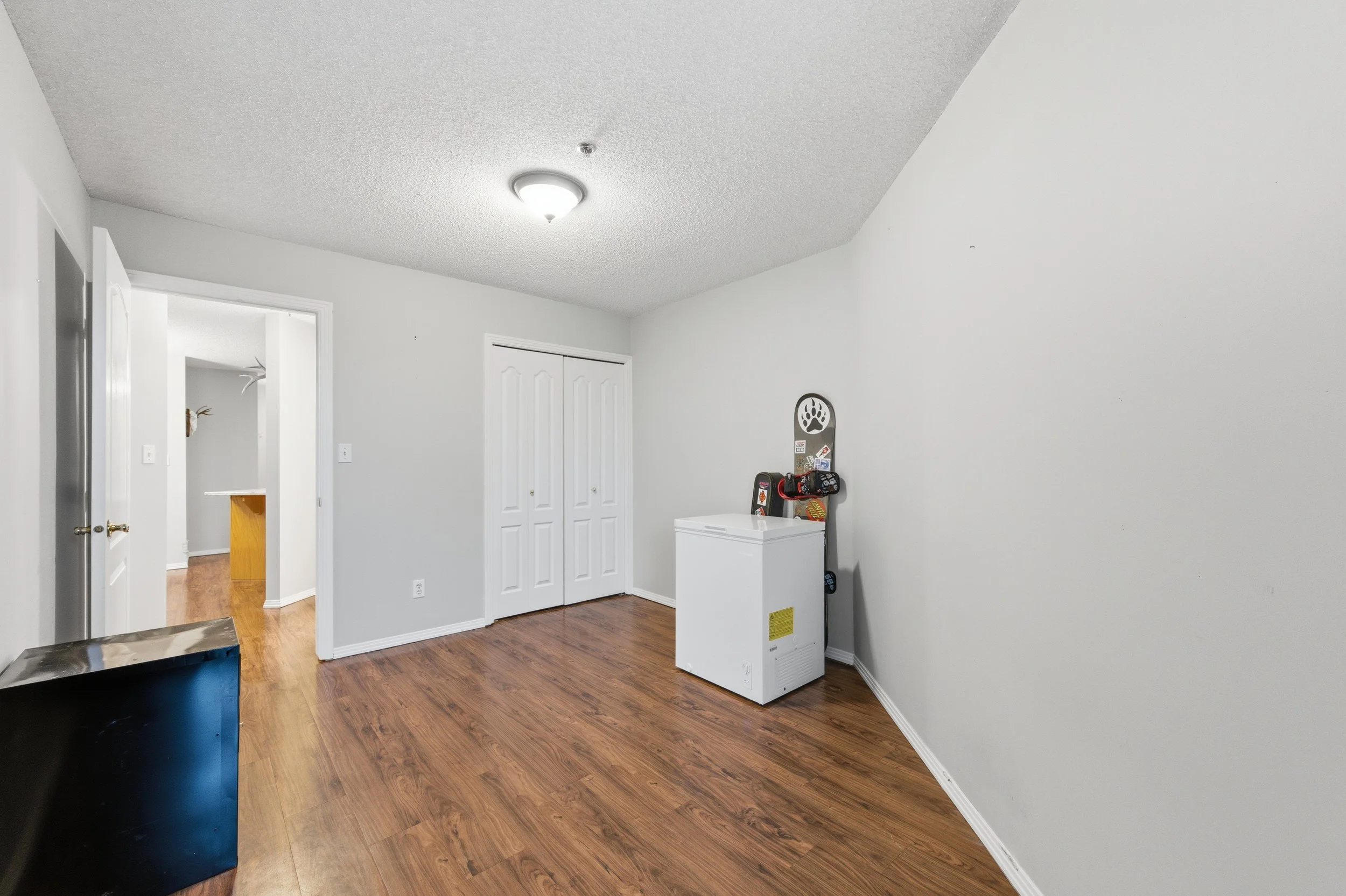 Empty room with wood flooring, white walls, a white ceiling light, a closet with double doors, a small black fridge, and a white chest freezer with a do-it-yourself kit hanging on the wall nearby.