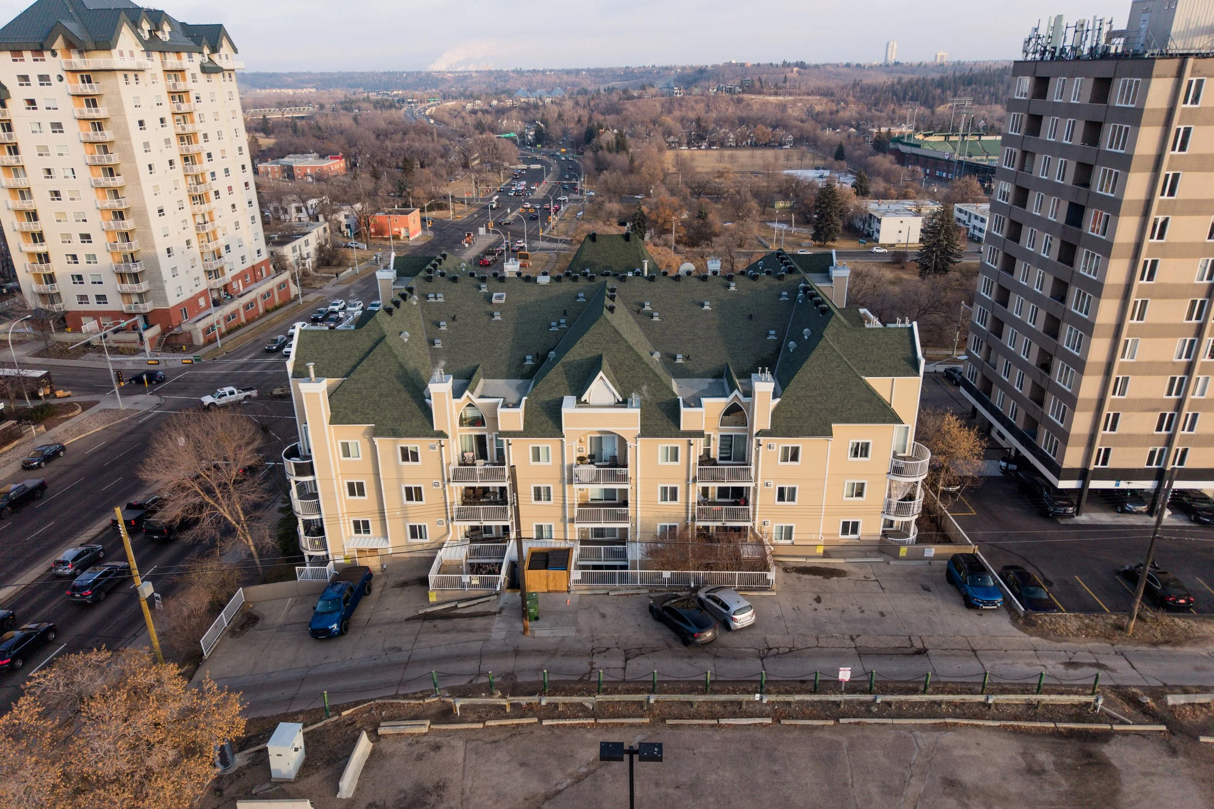 Aerial view of a yellow multi-story apartment building with green rooftops, surrounded by parking lots and nearby high-rise buildings in an urban area.