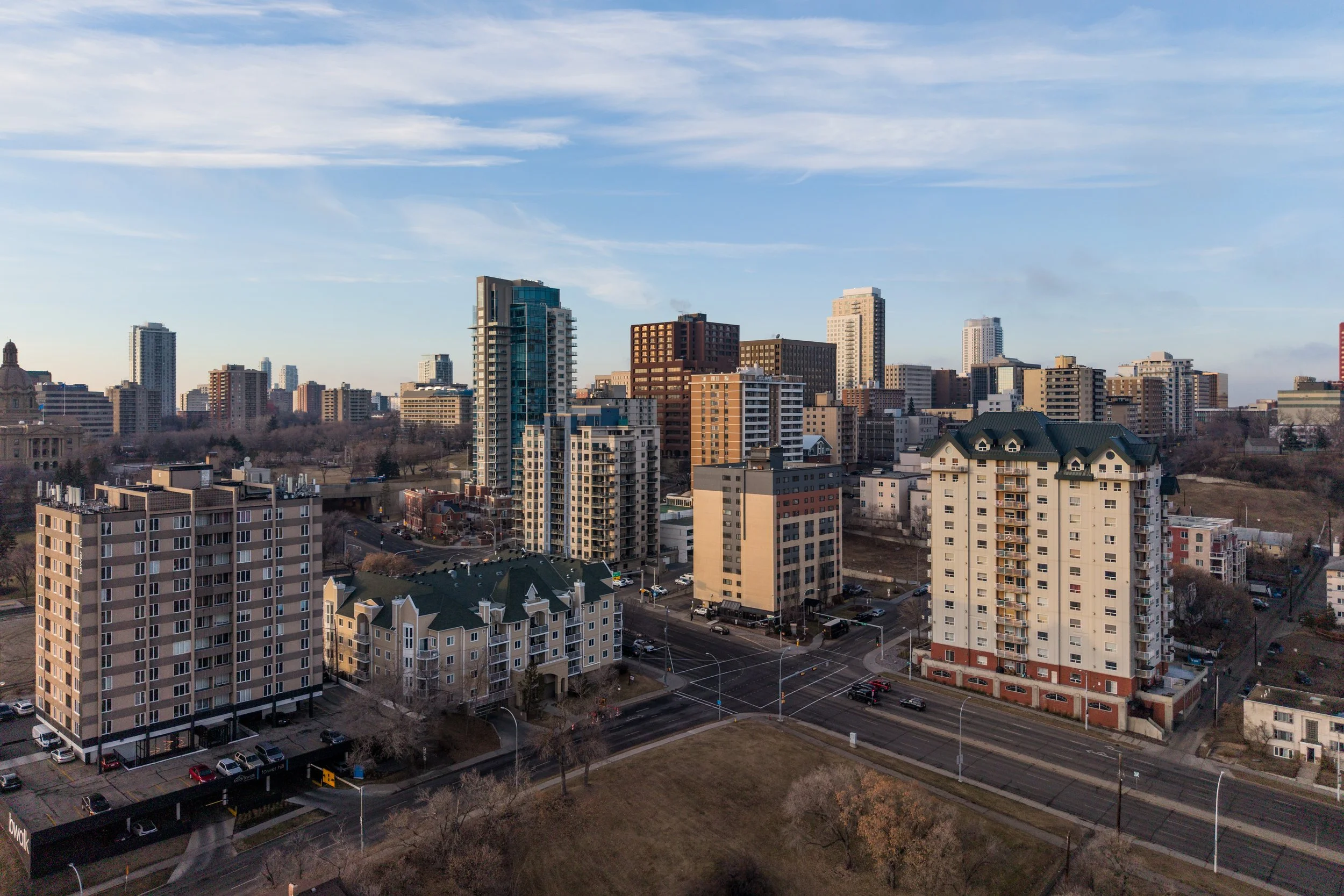 A cityscape with tall modern buildings, apartment complexes, and wide streets under a partly cloudy sky.