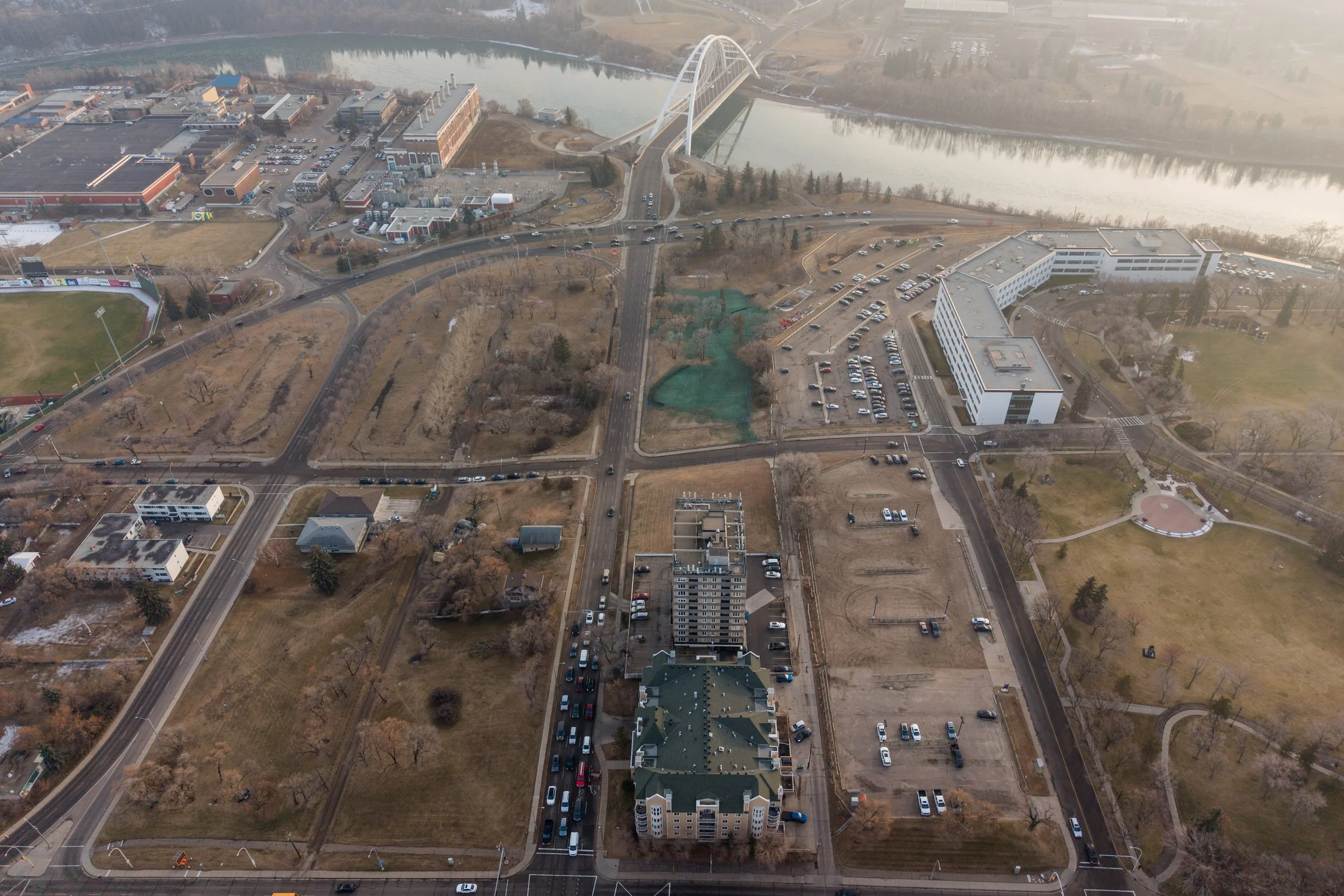 Aerial view of a cityscape with a river, several buildings, parking lots, roads, trees, and a bridge over the river.