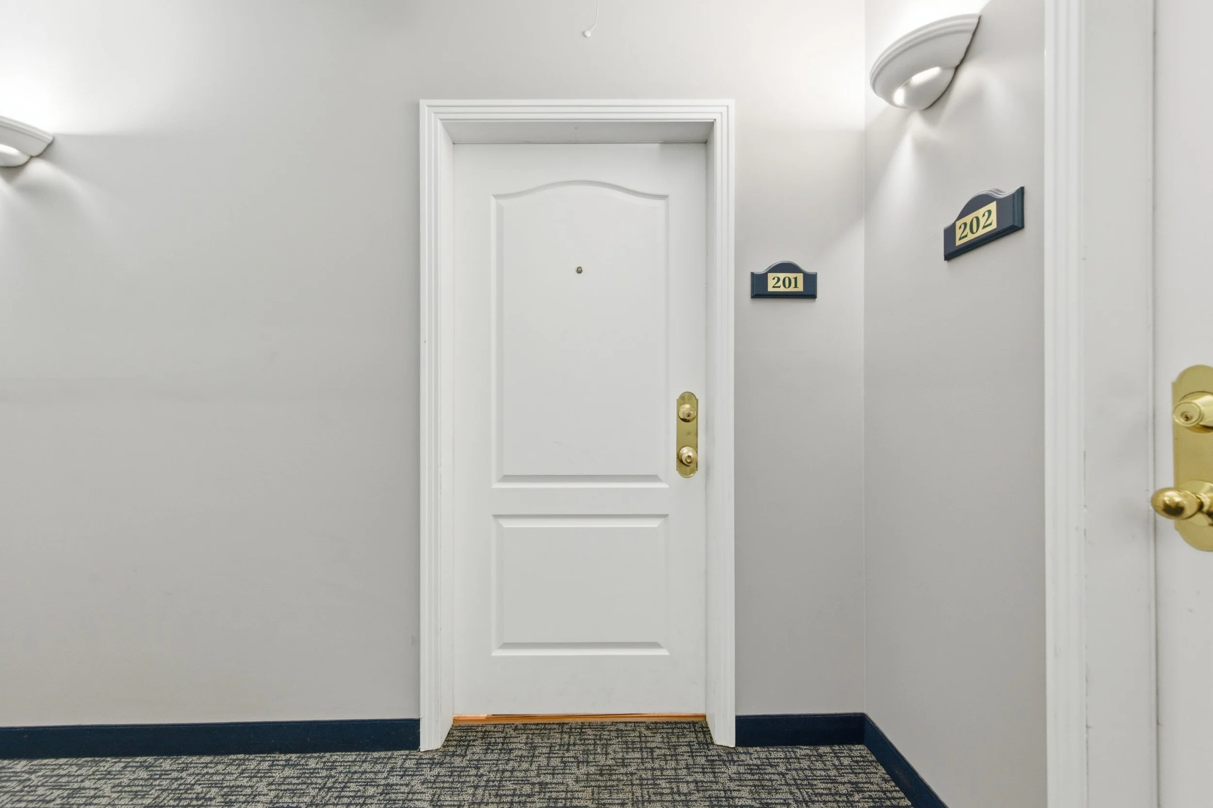 Interior view of an apartment building hallway with a closed white door, blue room number signs 201 and 202, and a beige carpeted floor.