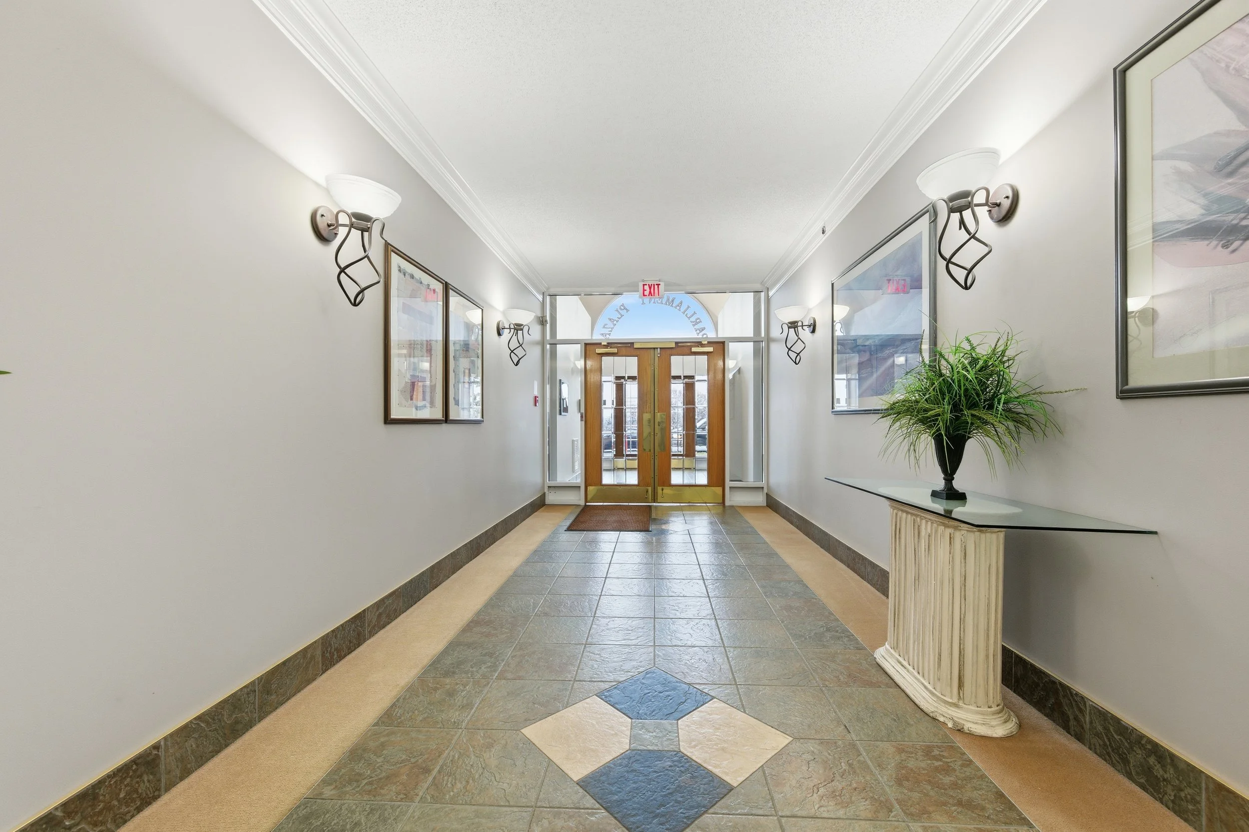 A hallway with light-colored walls, decorative wall sconces, framed artwork, a glass table with a plant, and wooden double doors at the end with an exit sign above.
