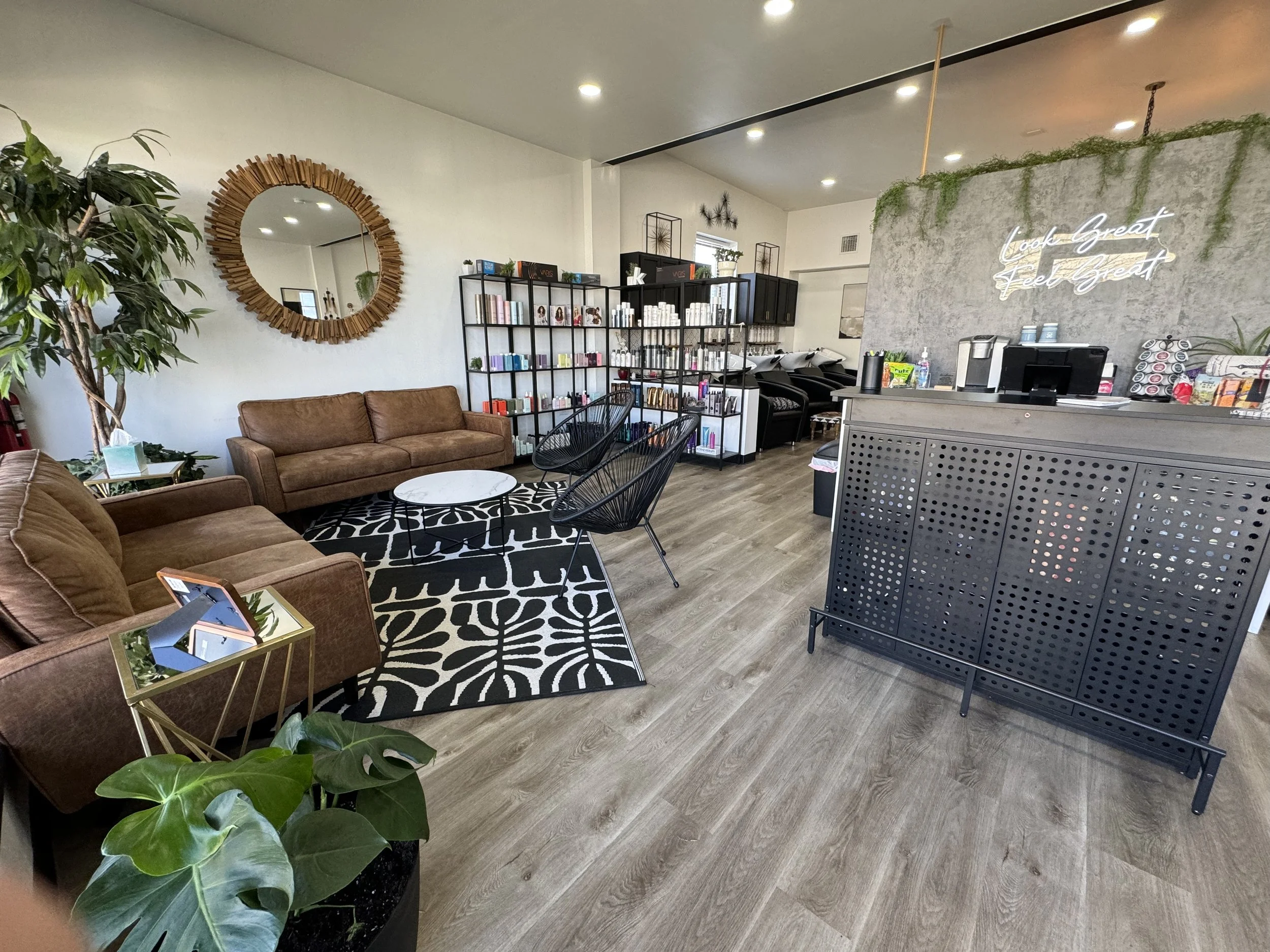 Interior of a modern salon with seating area including brown sofas, black chairs, and a patterned rug, shelves stocked with hair products, a round mirror, plants, and a coffee station with a neon sign on a textured wall.