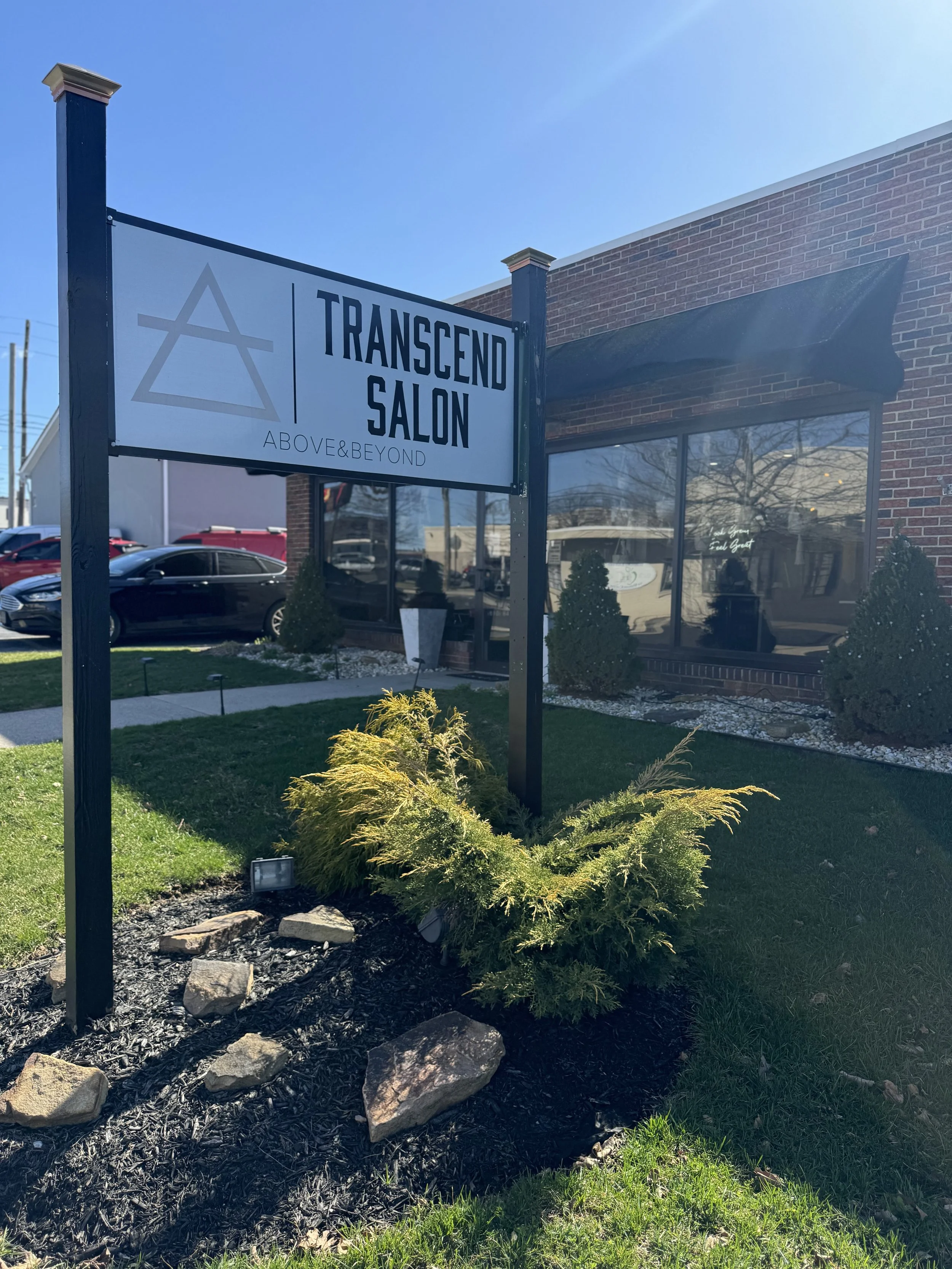 Sign for Transcend Salon with slogan 'Above & Beyond', located in front of a brick building, with small bushes and rocks in a landscaped area, and a parking lot with parked cars in the background.