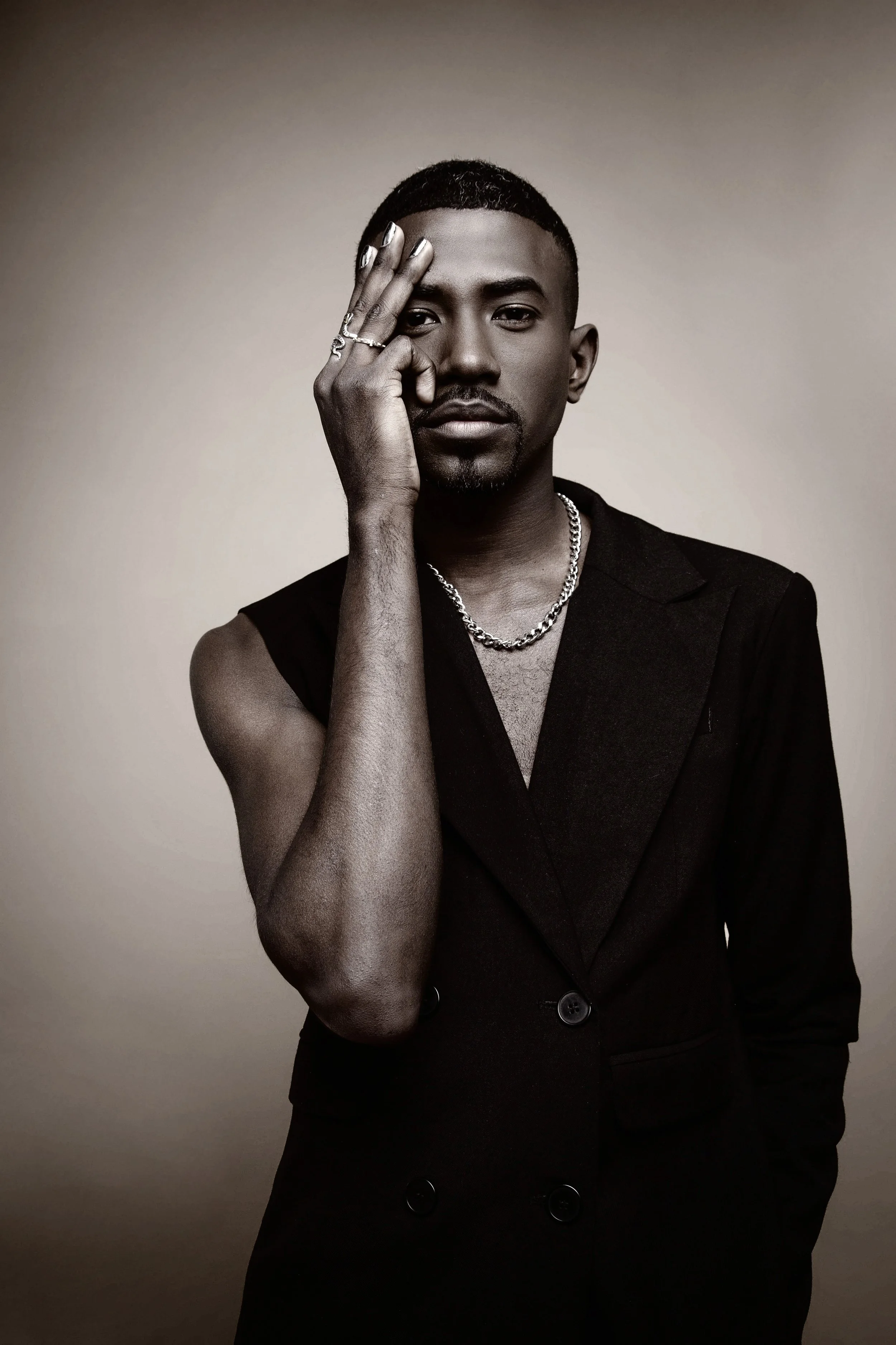 A black and white portrait of a man with a serious expression, wearing a sleeveless black blazer, a chain necklace, and rings, with his hand partially covering one eye against a plain background.