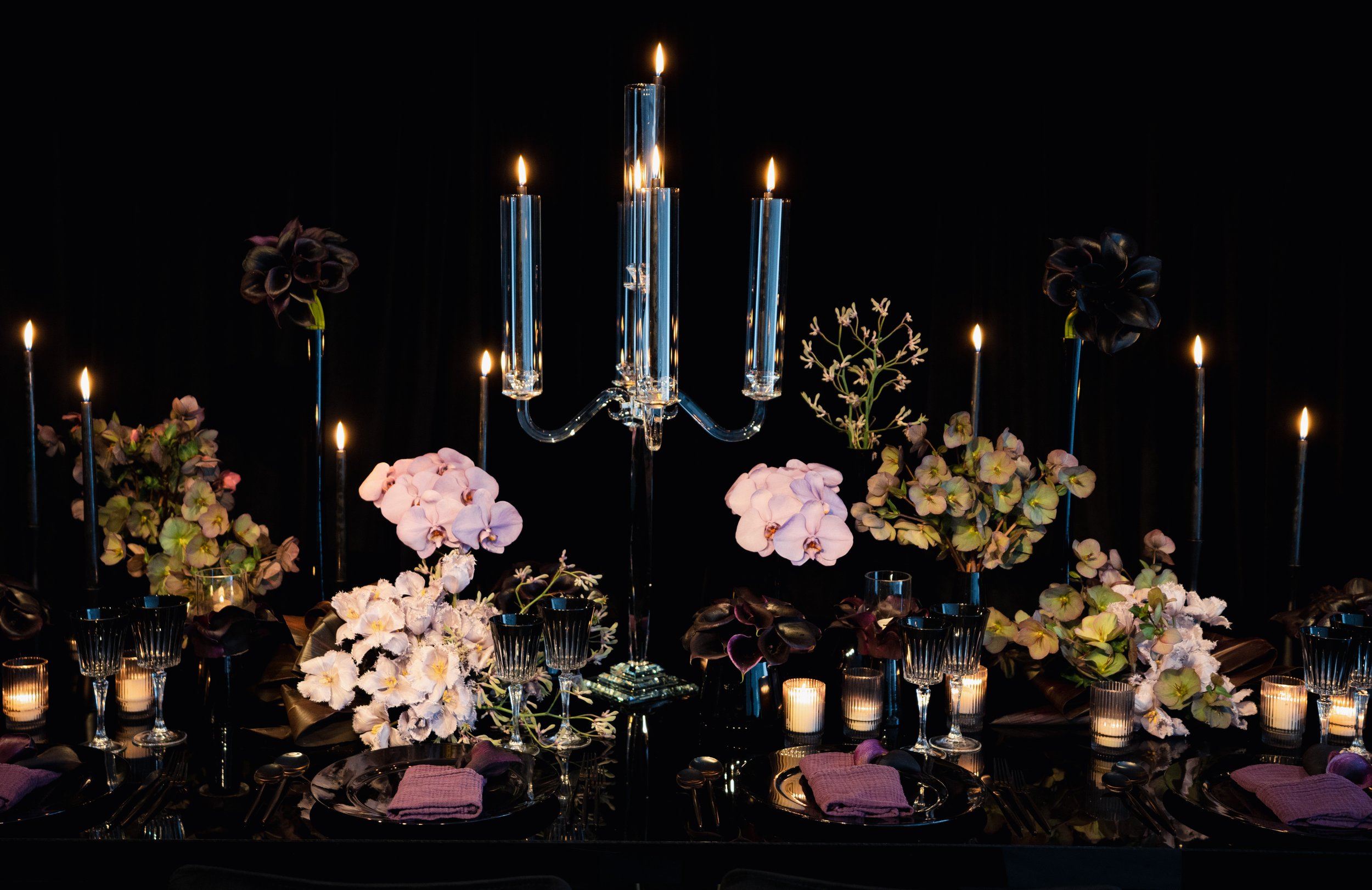 Elegant dinner table setting with floral arrangements, candles, and a large glass candelabrum with five lit candles.